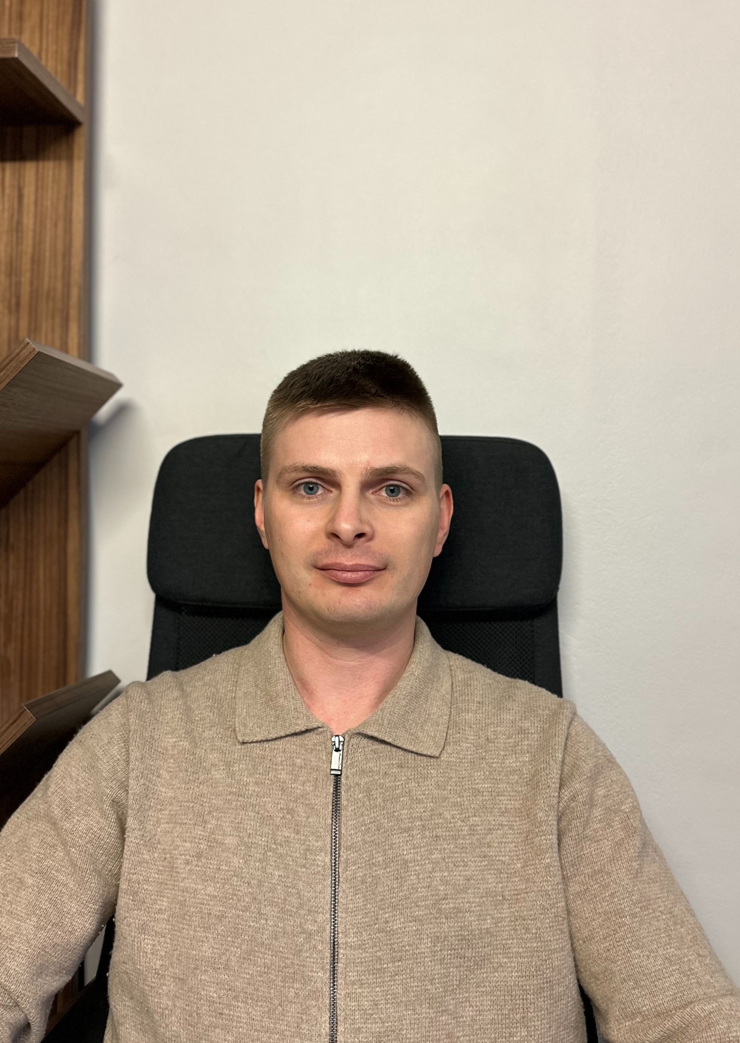 Portret mężczyzny w beżowym swetrze z zamkiem błyskawicznym, siedzącego na czarnym krześle biurowym na tle białej ściany i fragmentu drewnianego regału.