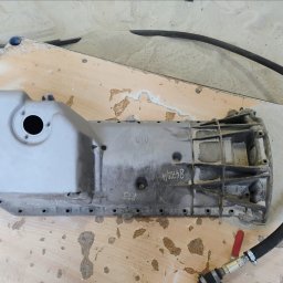 Pablopiask .Piaskowanie - Szara, aluminiowa miska olejowa BMW, częściowo oczyszczona, z widocznymi numerami '8472/4' i 'R73', leżąca na drewnianej powierzchni z resztkami piasku, obok czarny wąż z czerwonym zaworem.