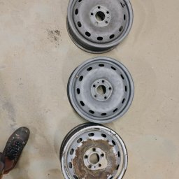 Pablopiask .Piaskowanie - Trzy stalowe felgi samochodowe ułożone jedna pod drugą na betonowej posadzce, widoczna stopa w roboczym obuwiu w lewym dolnym rogu, felga na dole z widocznymi śladami korozji przed obróbką.