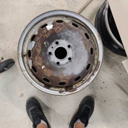 Pablopiask .Piaskowanie - Oczyszczona z rdzy felga samochodowa z widocznymi śladami korozji i resztkami farby, częściowo pomalowana szarą farbą podkładową, sfotografowana z góry na betonowej posadzce garażu, z butami osoby...