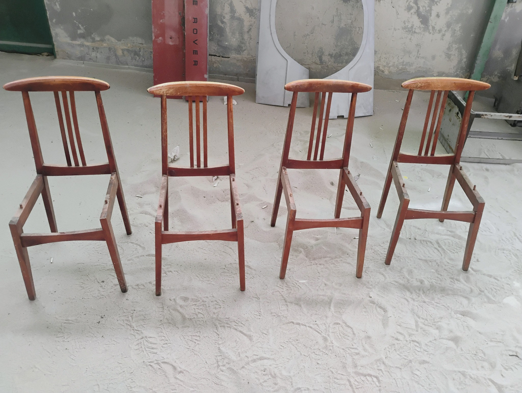 Cztery drewniane krzesła bez siedzisk, ustawione w rzędzie w pomieszczeniu z betonową podłogą pokrytą pyłem, prawdopodobnie po piaskowaniu lub innym procesie czyszczenia.