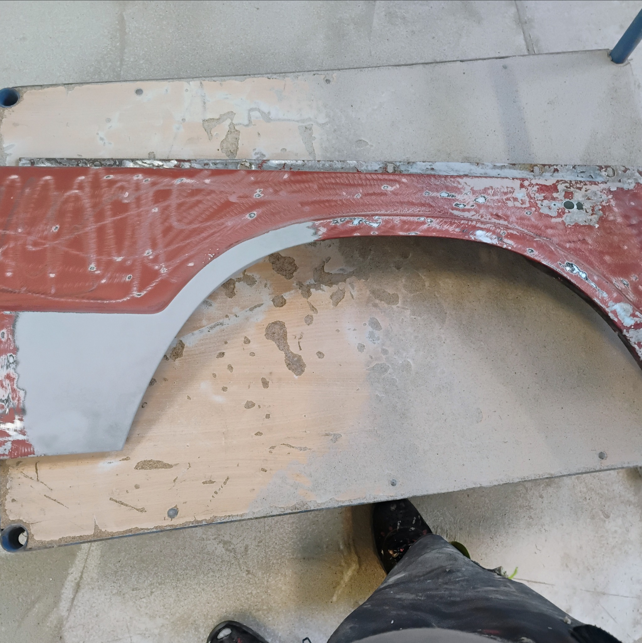 Fragment karoserii samochodowej, wstępnie przygotowany do lakierowania, z widocznymi śladami szlifowania i nałożonym podkładem, leżący na warsztatowym stole. W rogu kadru widoczny fragment ubrania...