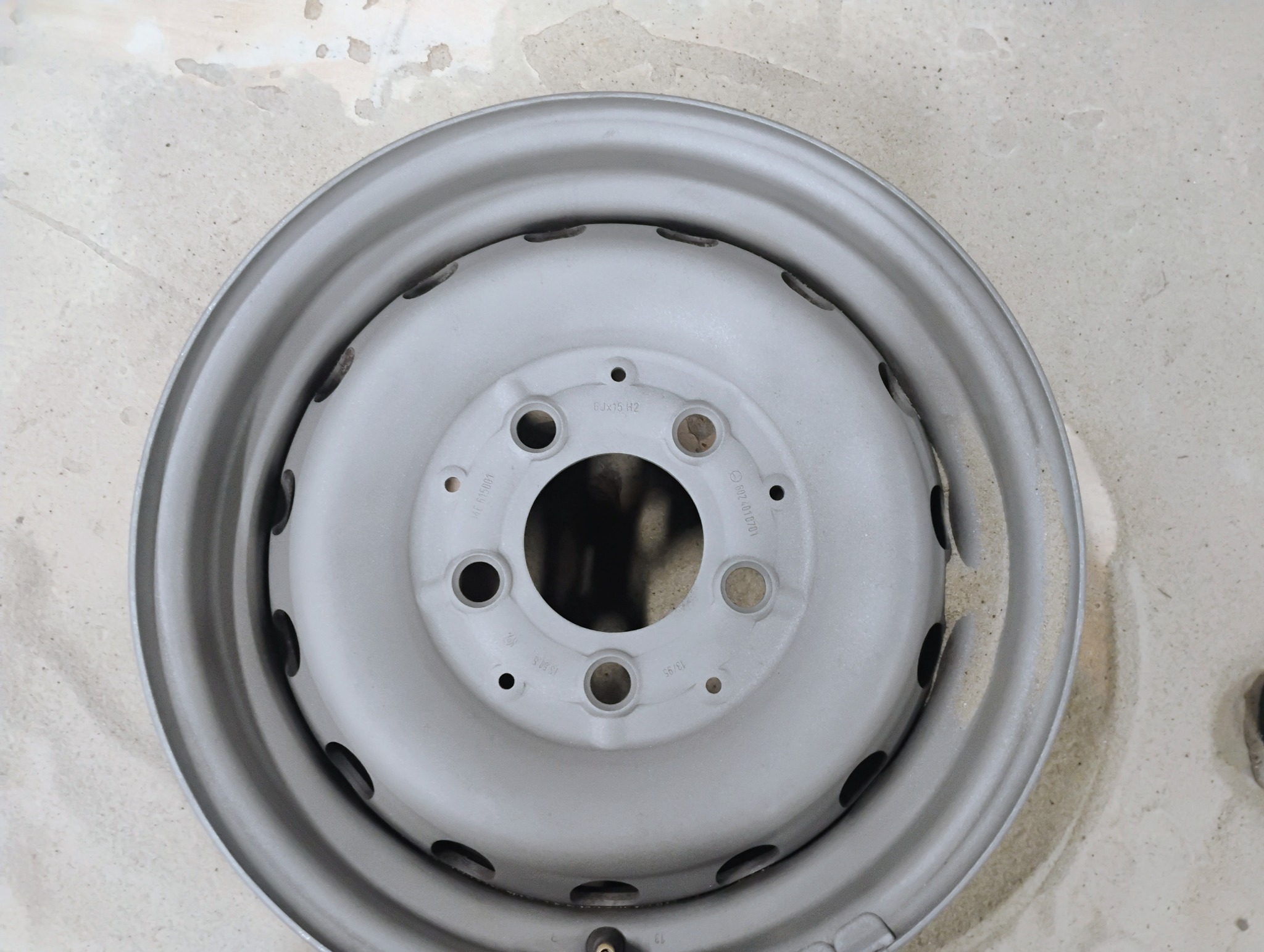 Pomalowana na szaro felga samochodowa na tle betonowej posadzki, widoczne otwory na śruby i wentyl.