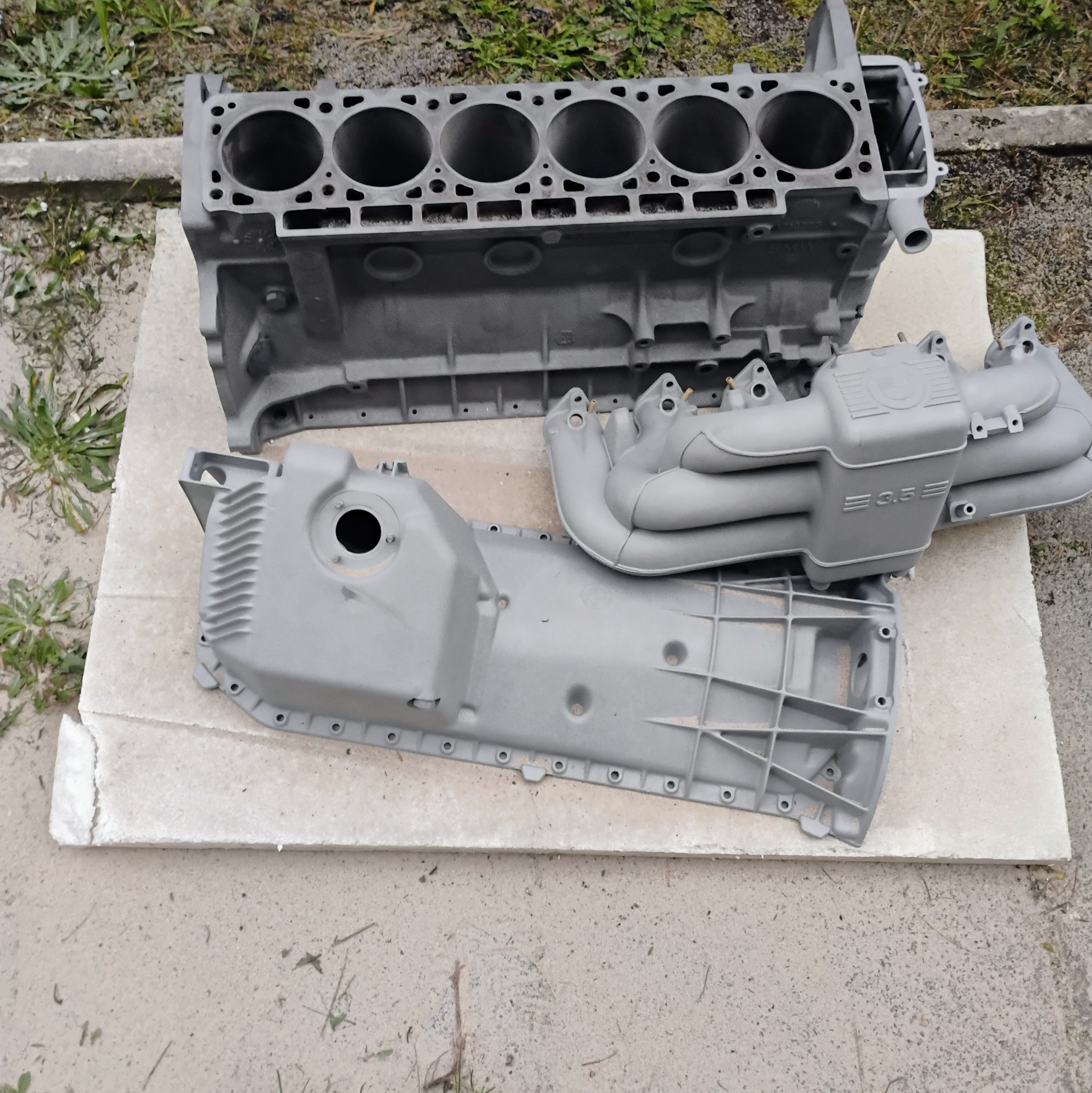 Szary, odnowiony blok silnika BMW, kolektor ssący i miska olejowa leżące na betonowej płycie na zewnątrz.