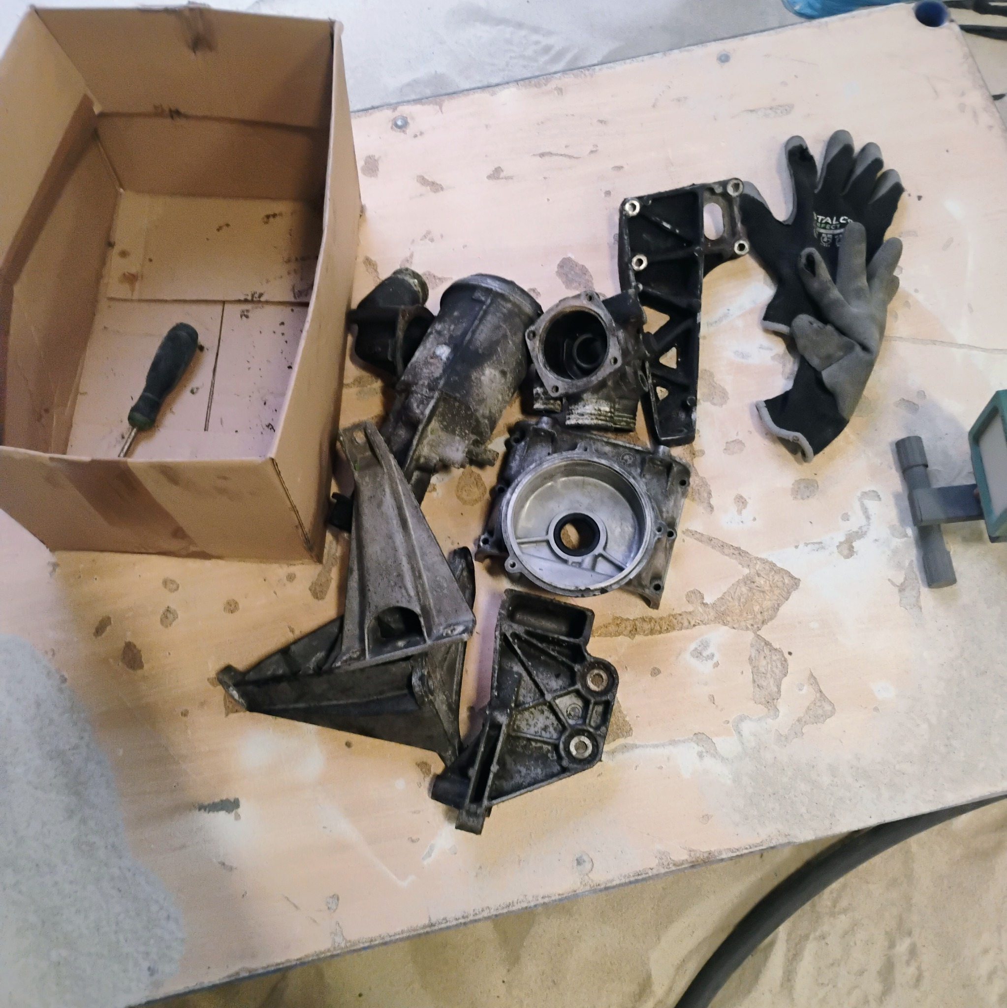 Zdemontowane, brudne elementy silnika samochodowego, leżące na stole obok kartonowego pudełka z wkrętakiem i rękawic roboczych.
