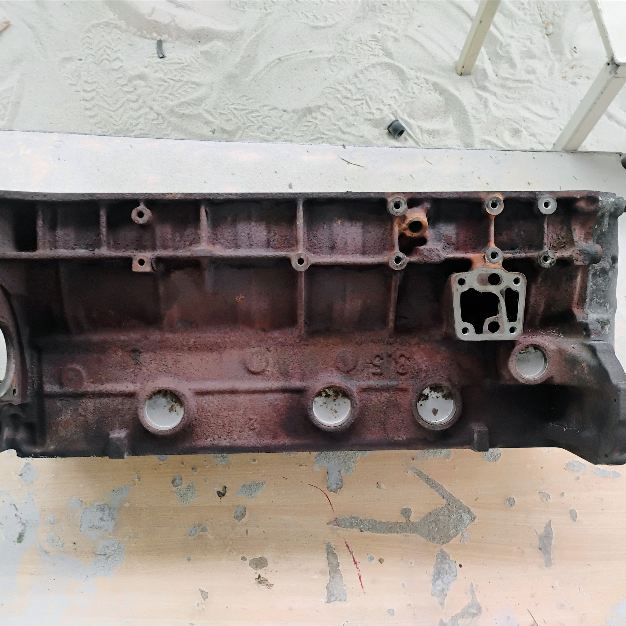 Zardzewiały blok silnika spalinowego, widoczne ślady korozji i otwory montażowe, leżący na powierzchni z odpryskami farby.