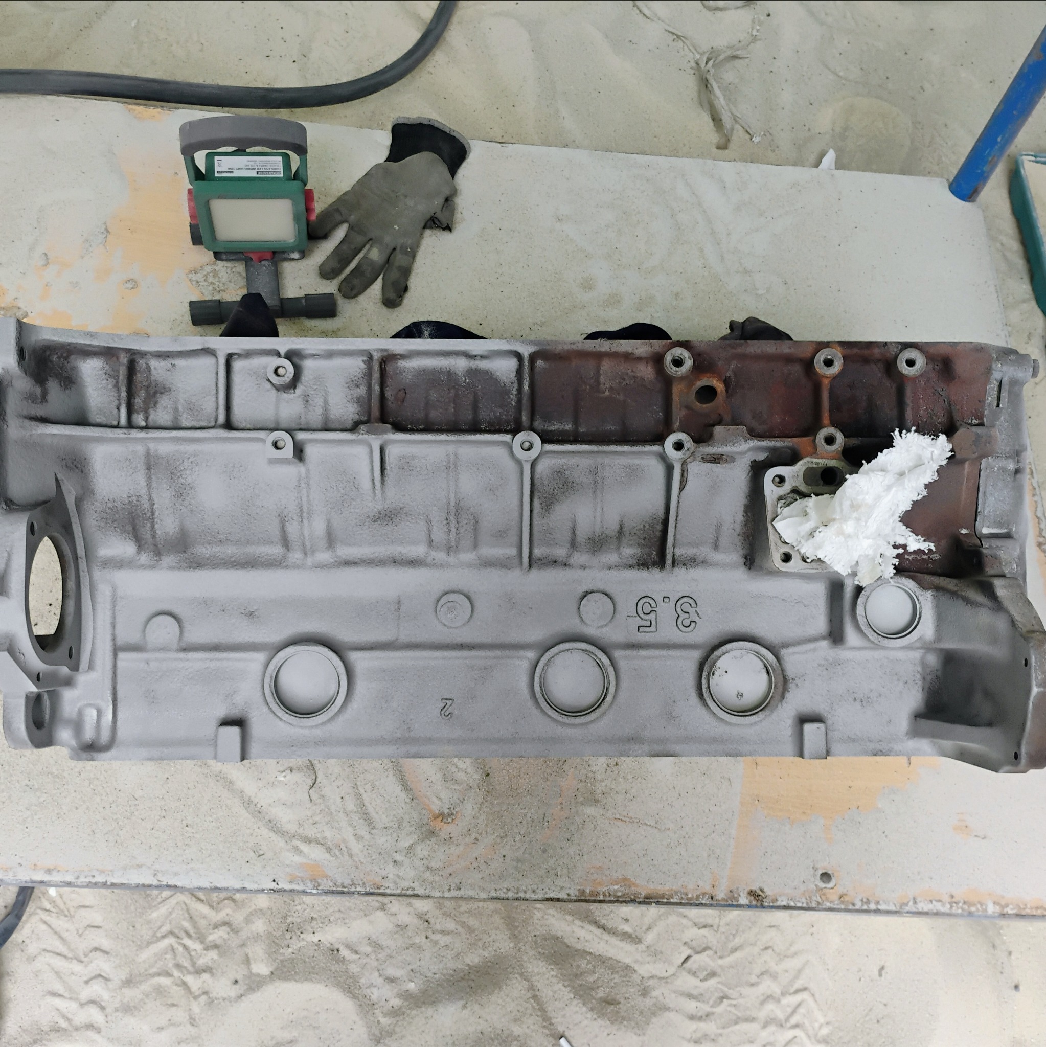 Oczyszczony blok silnika 3.5, częściowo pomalowany szarą farbą, leżący na piasku obok lampy inspekcyjnej i rękawicy roboczej.