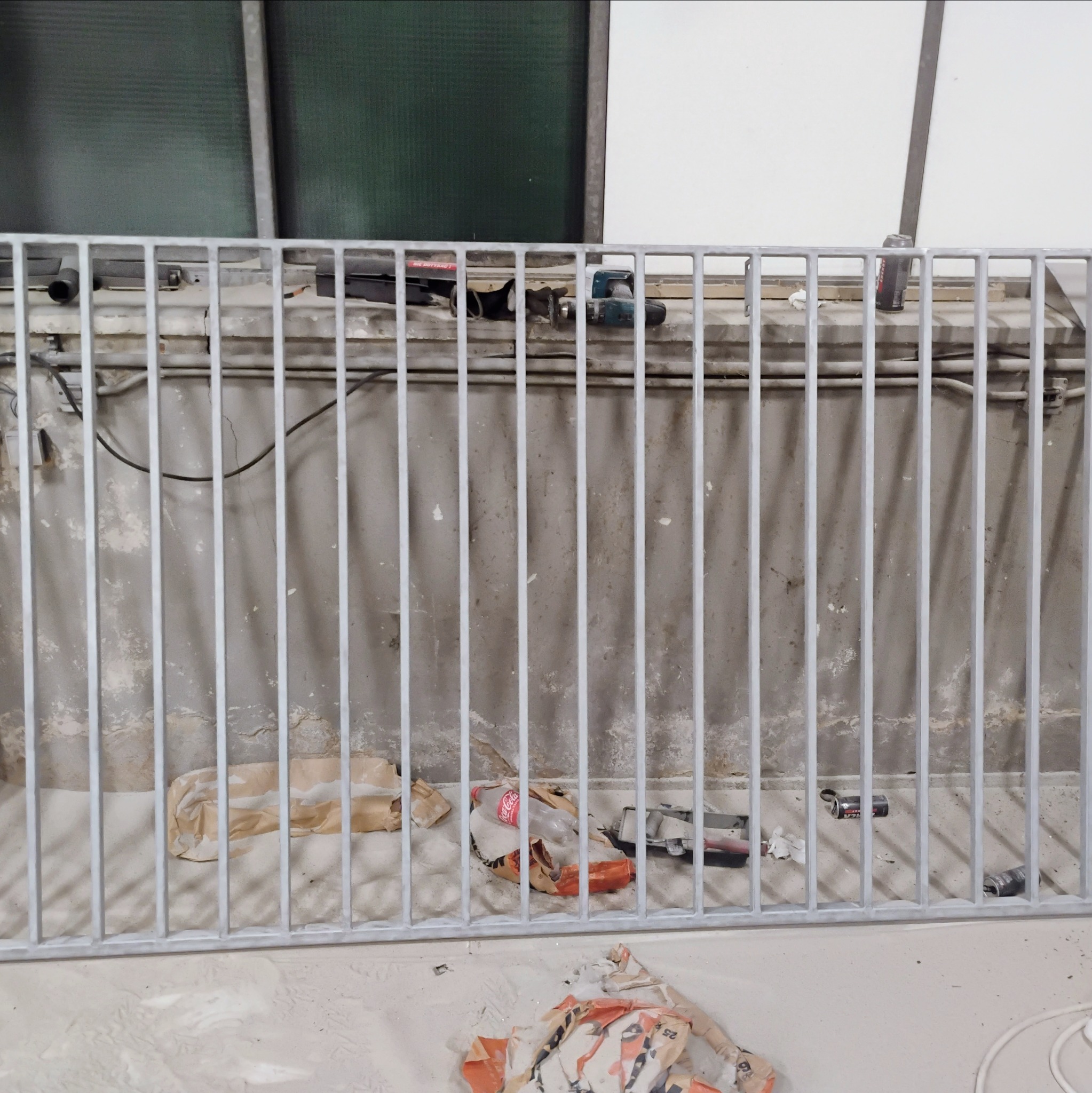 Szara, metalowa balustrada po malowaniu, widoczne ślady farby na podłodze i ścianie, puszki po napojach i narzędzia malarskie w tle.