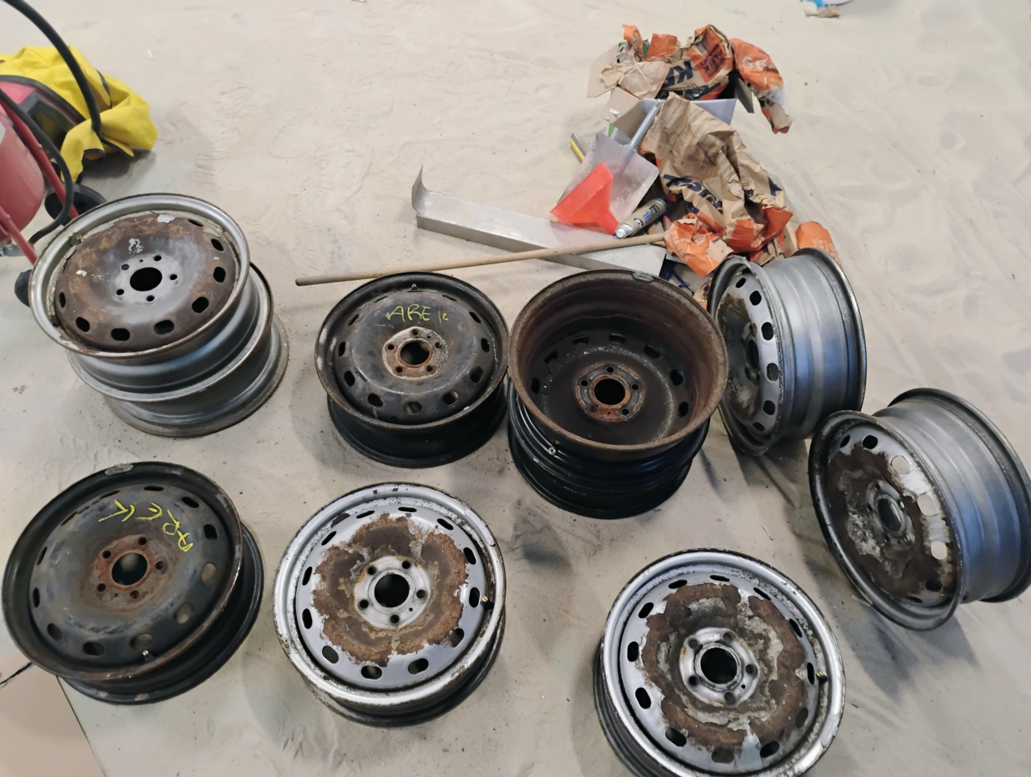 Stare, zardzewiałe felgi samochodowe przed renowacją, z widocznymi śladami korozji i żółtym napisem na jednej z nich, leżące na betonowej podłodze w warsztacie.