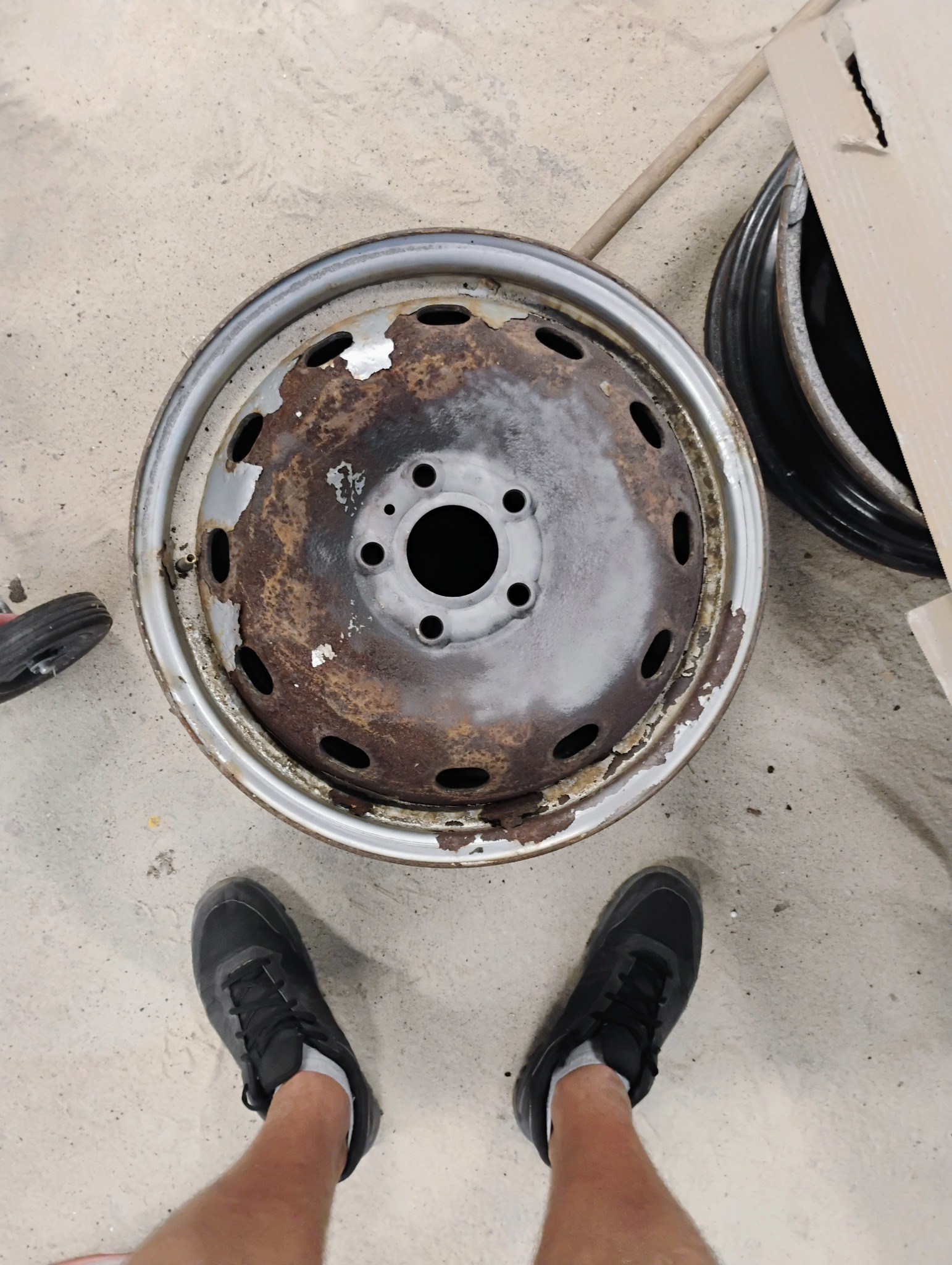 Oczyszczona z rdzy felga samochodowa z widocznymi śladami korozji i resztkami farby, częściowo pomalowana szarą farbą podkładową, sfotografowana z góry na betonowej posadzce garażu, z butami osoby...