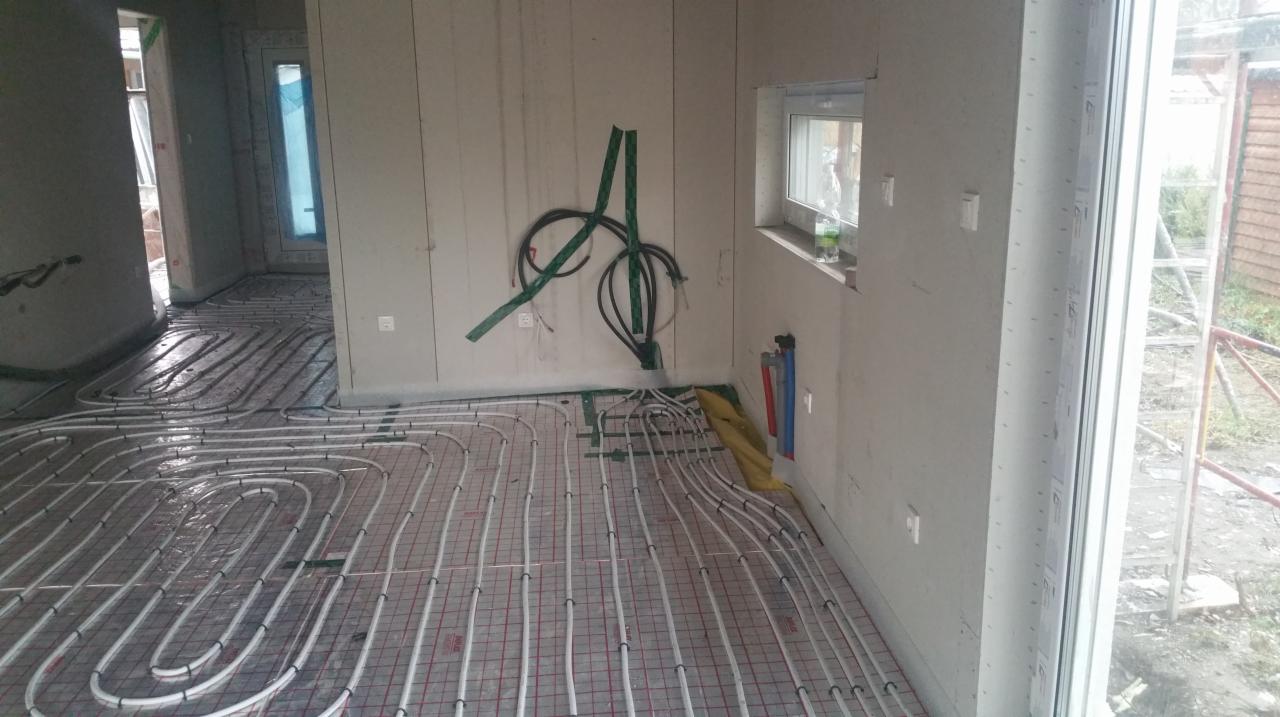 Wnętrze w trakcie instalacji ogrzewania podłogowego, rury grzewcze rozłożone na siatce, widoczne przyłącza i kable wystające ze ściany. Okno z widokiem na zewnątrz.