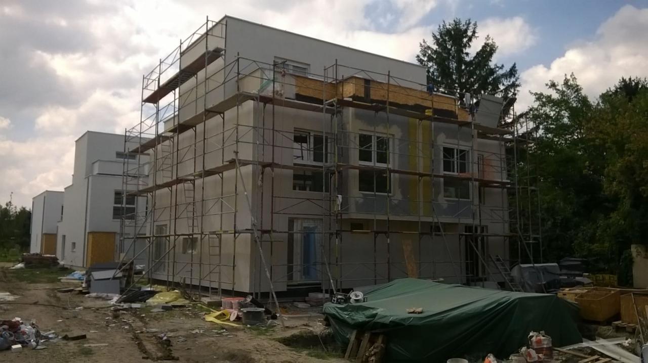 Wielorodzinny budynek w trakcie budowy, z widocznymi rusztowaniami, ociepleniem i elementami konstrukcyjnymi, teren budowy z gruzem i przykrytymi materiałami.