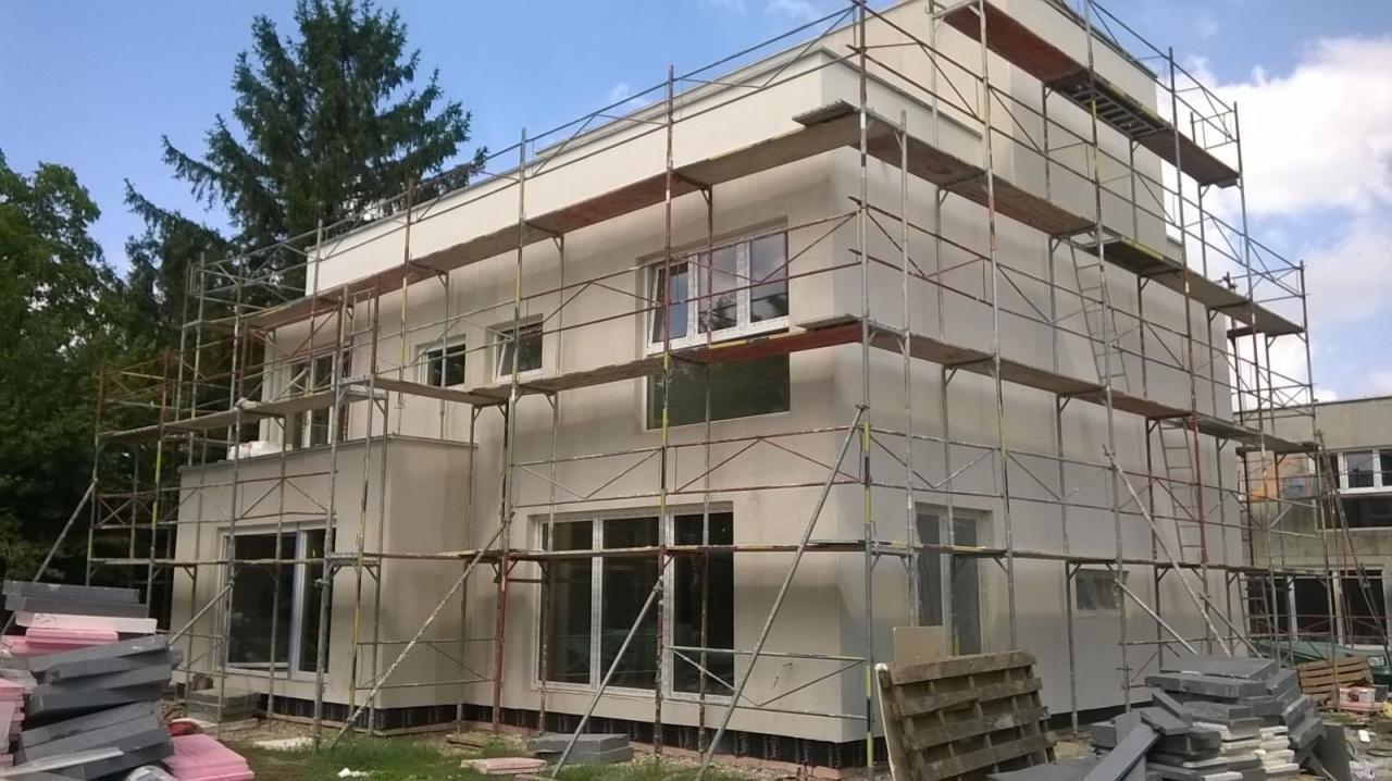Dwukondygnacyjny budynek w trakcie prac wykończeniowych elewacji, otoczony rusztowaniem, z widocznymi materiałami izolacyjnymi i wykończeniowymi na pierwszym planie.