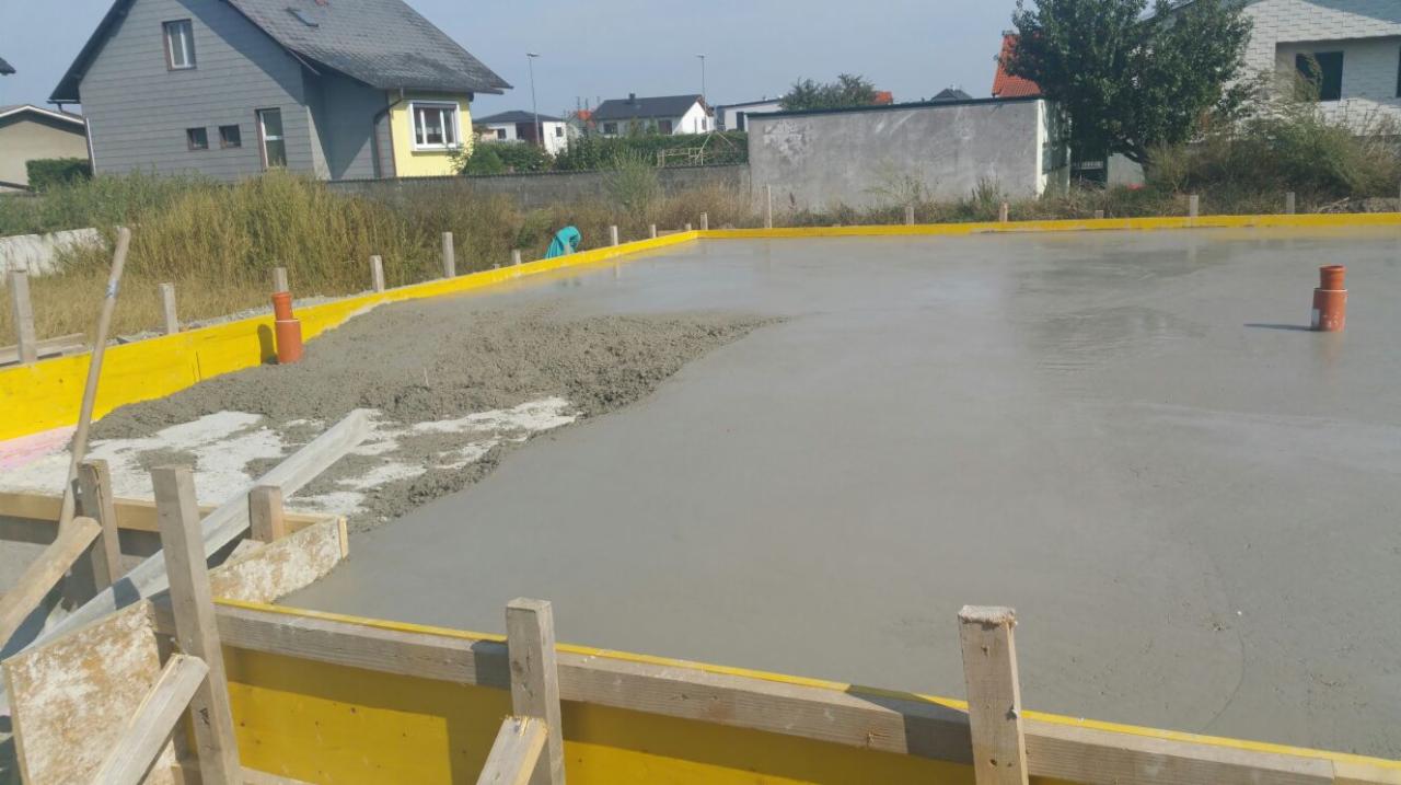 Wylewka betonowa na przygotowanym fundamencie z żółtymi szalunkami, widoczne rury instalacyjne i częściowo wyrównany beton, budowa w toku.