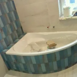 Narożna wanna obudowana niebiesko-szarymi płytkami w trakcie montażu, widoczne narzędzia i materiały budowlane na parapecie okna.