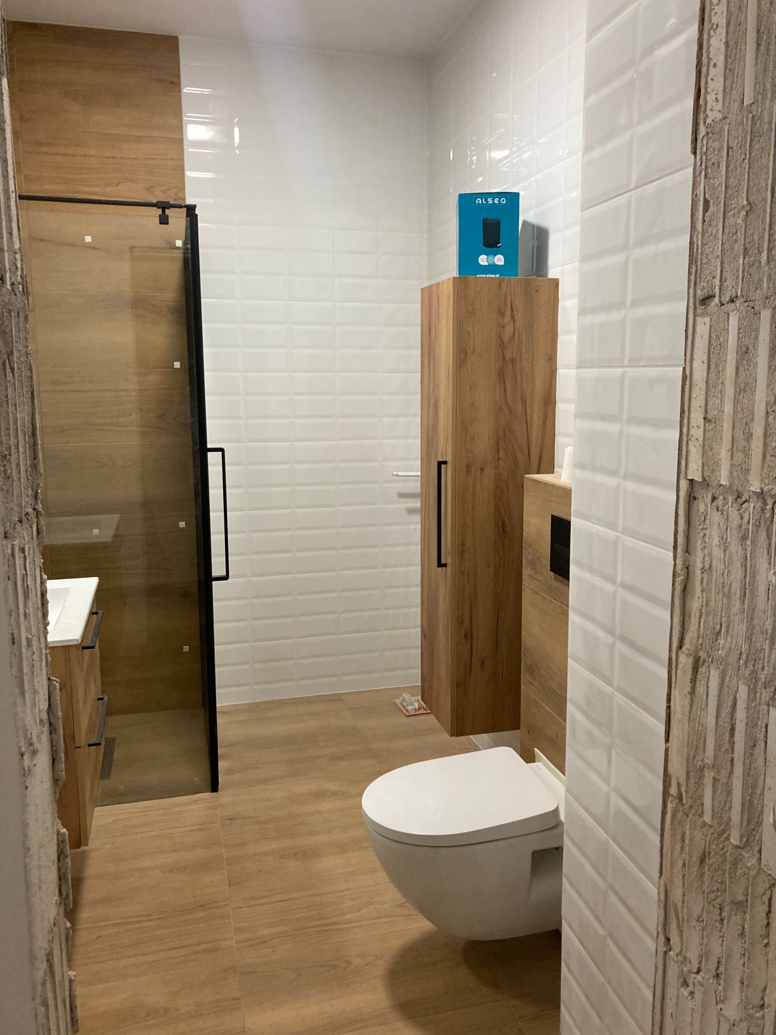 Nowoczesna łazienka z kabiną prysznicową ze szkła, drewnianą szafką, toaletą i białymi płytkami imitującymi cegłę, ściana w stanie surowym.