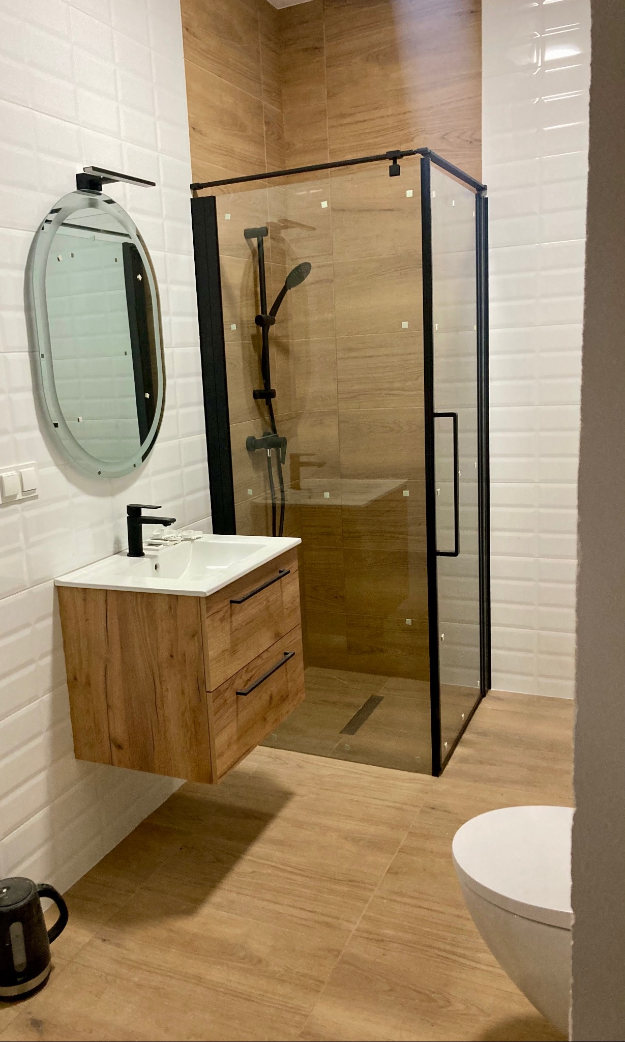 Nowoczesna łazienka z drewnopodobnymi płytkami, kabiną prysznicową z czarnymi profilami, wiszącą szafką z umywalką i owalnym lustrem z podświetleniem