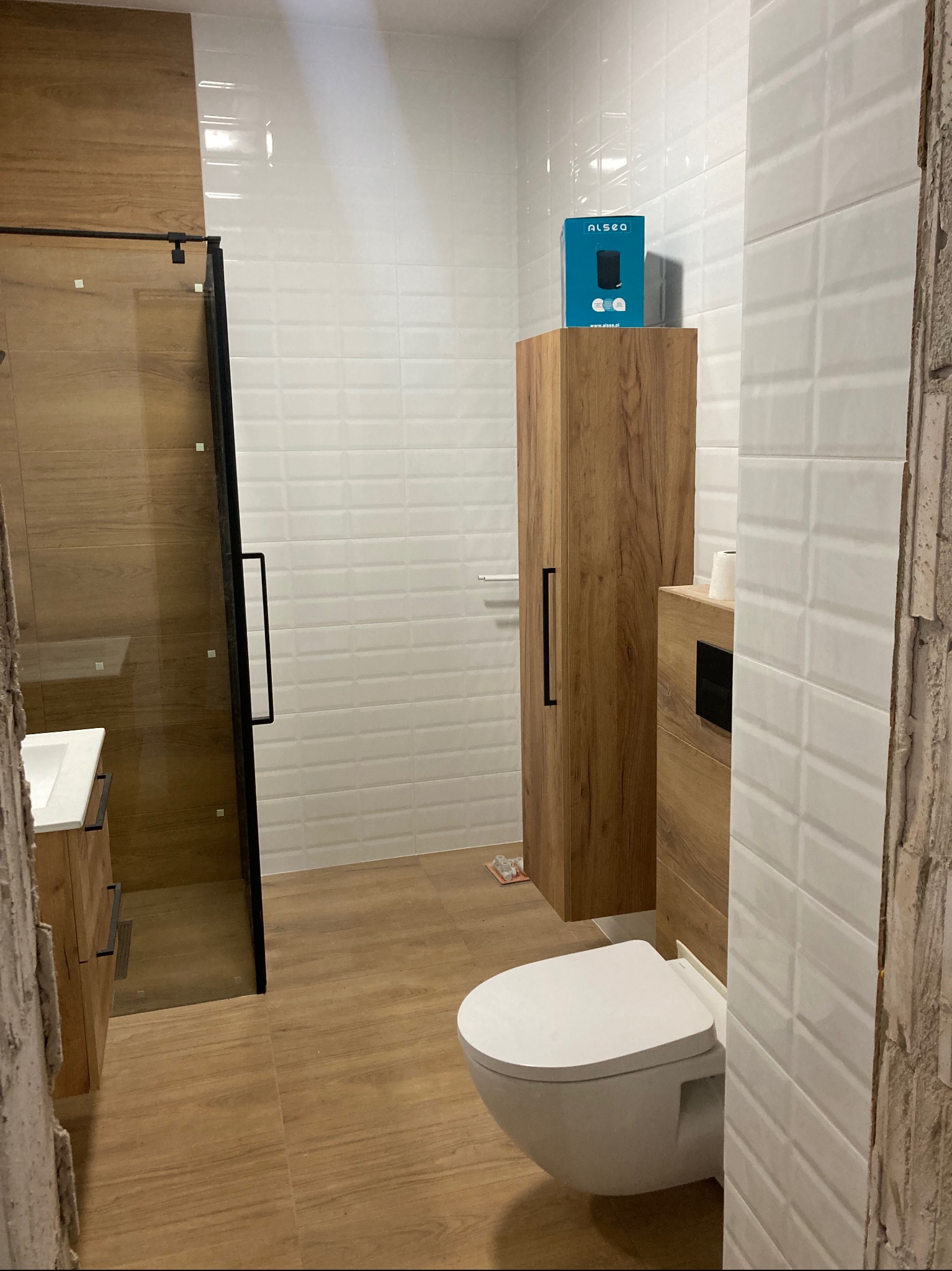 Nowoczesna łazienka z kabiną prysznicową ze szklanymi drzwiami w czarnej ramie, wisząca toaleta, drewniana szafka z czarnym uchwytem oraz białe płytki na ścianach. Na szafce stoi niebieskie pudełko...