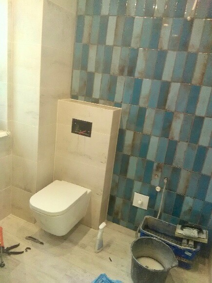 Montaż podwieszanej toalety w łazience z niebiesko-szarymi płytkami, widoczne narzędzia i pojemnik z zaprawą.