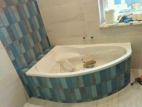 Narożna wanna obudowana niebiesko-szarymi płytkami w trakcie montażu, widoczne narzędzia i materiały budowlane na parapecie okna.