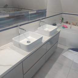 KOMPLEKSOWE USŁUGI HYDRAULICZNE ADRIAN WESOŁOWSKI - Nowoczesna łazienka na poddaszu z dwiema kwadratowymi umywalkami nablatowymi, chromowanymi bateriami i szarą szafką pod umywalką. W tle widoczna wanna i akcesoria łazienkowe.