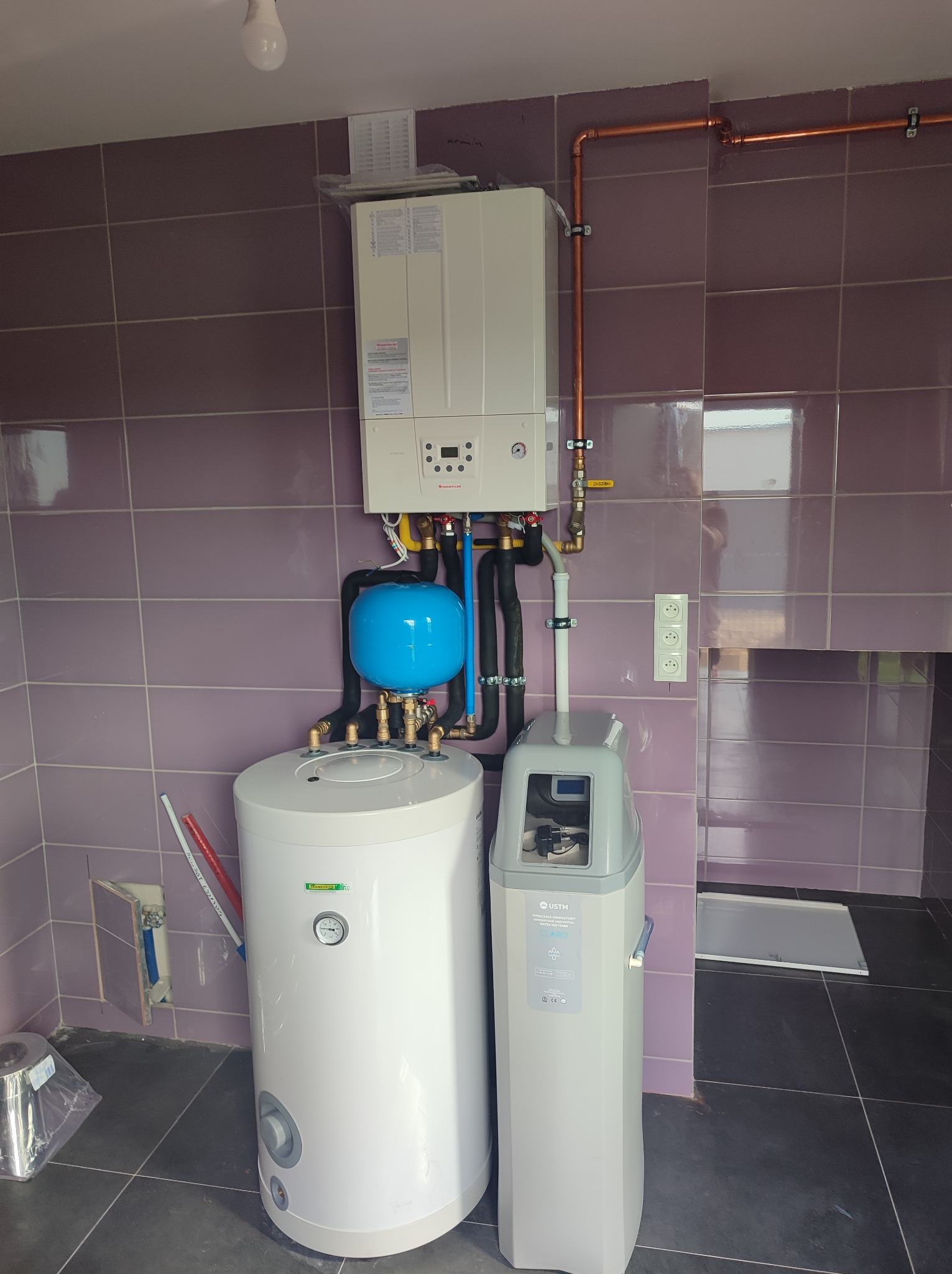 Nowoczesna kotłownia w fioletowej glazurze: biały piec gazowy wiszący nad bojlerem z niebieskim naczyniem przeponowym, obok zmiękczacz wody. Miedziane rury instalacji grzewczej biegną wzdłuż sufitu.
