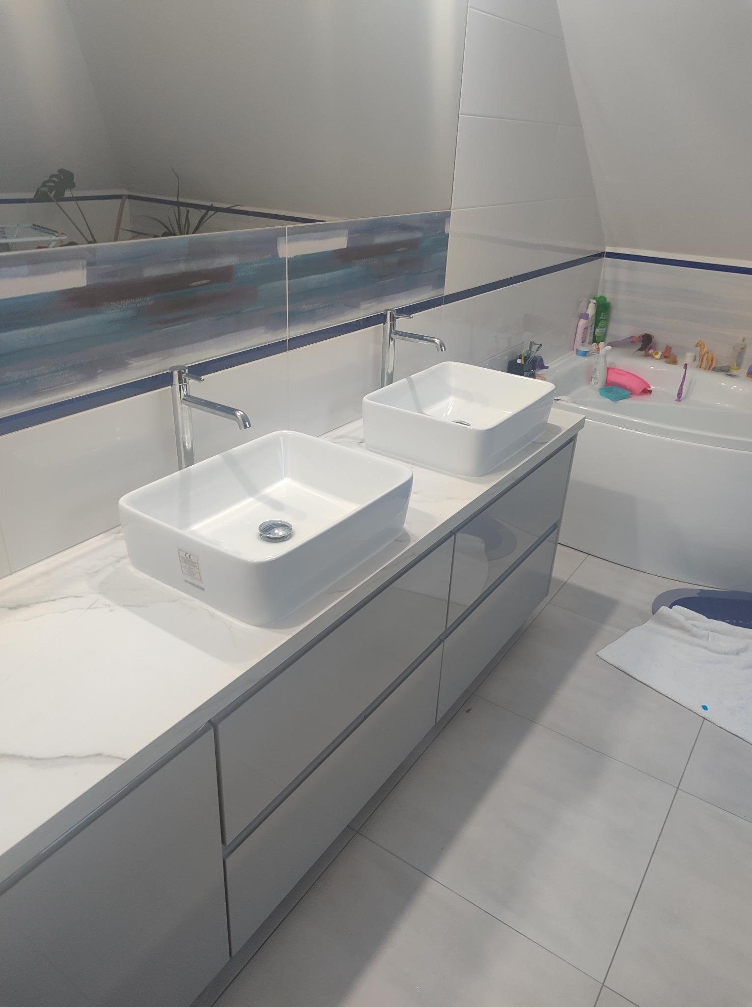 Nowoczesna łazienka na poddaszu z dwiema kwadratowymi umywalkami nablatowymi, chromowanymi bateriami i szarą szafką pod umywalką. W tle widoczna wanna i akcesoria łazienkowe.