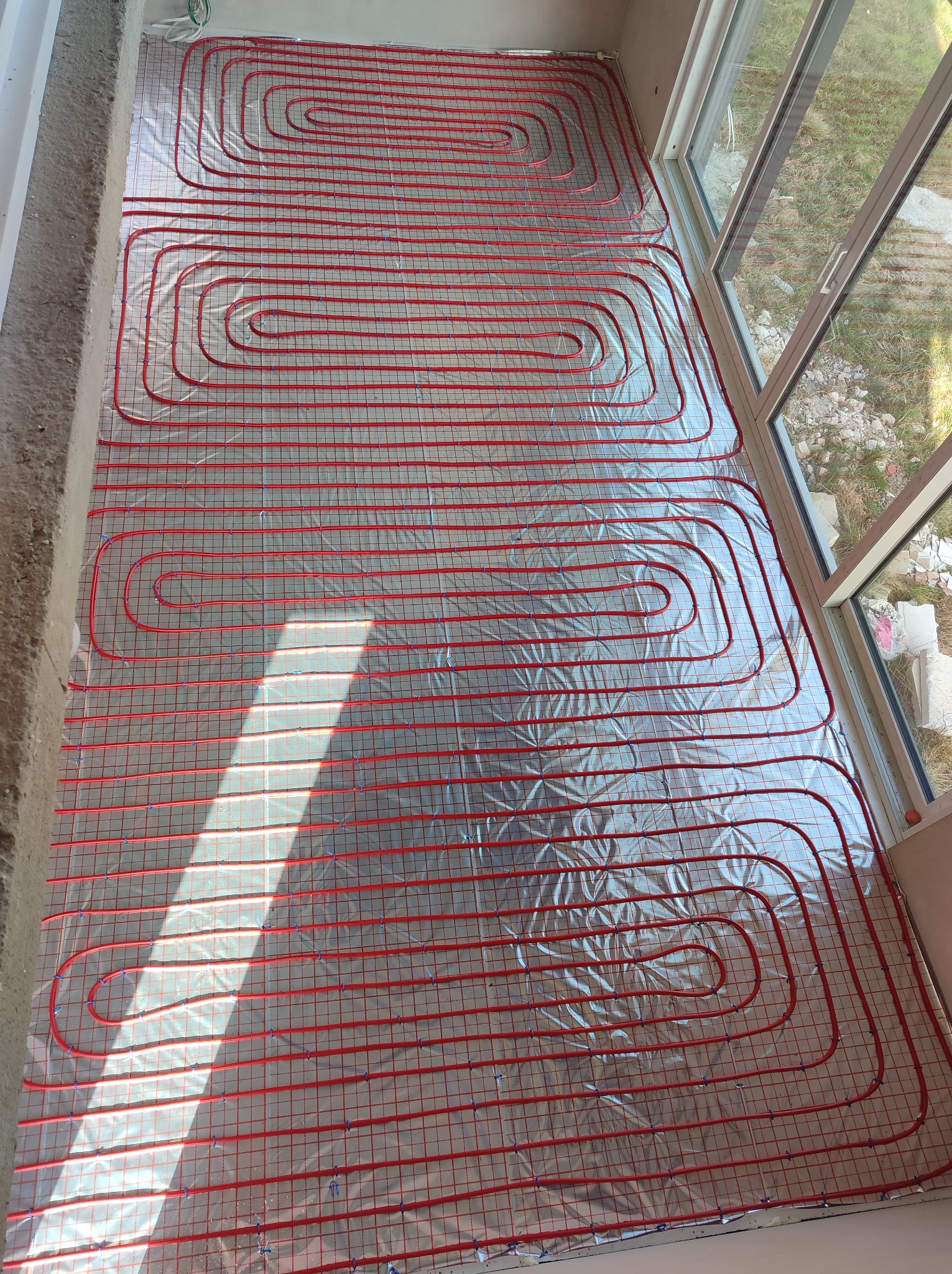 Czerwone rurki ogrzewania podłogowego ułożone na srebrnej folii izolacyjnej w nowym budynku, widoczne okno z zewnątrz.