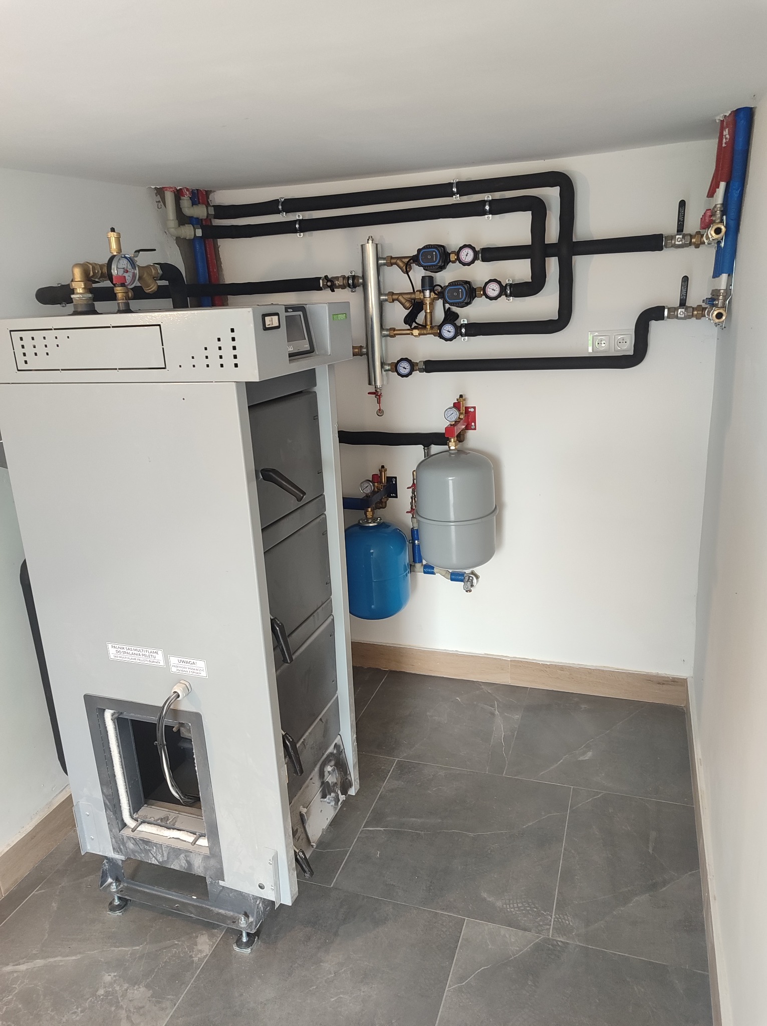 Kocioł na paliwo stałe w kotłowni, z widoczną instalacją grzewczą z czarnymi rurami, manometrami i zbiornikami wyrównawczymi, na tle białych ścian i szarej podłogi z płytek.
