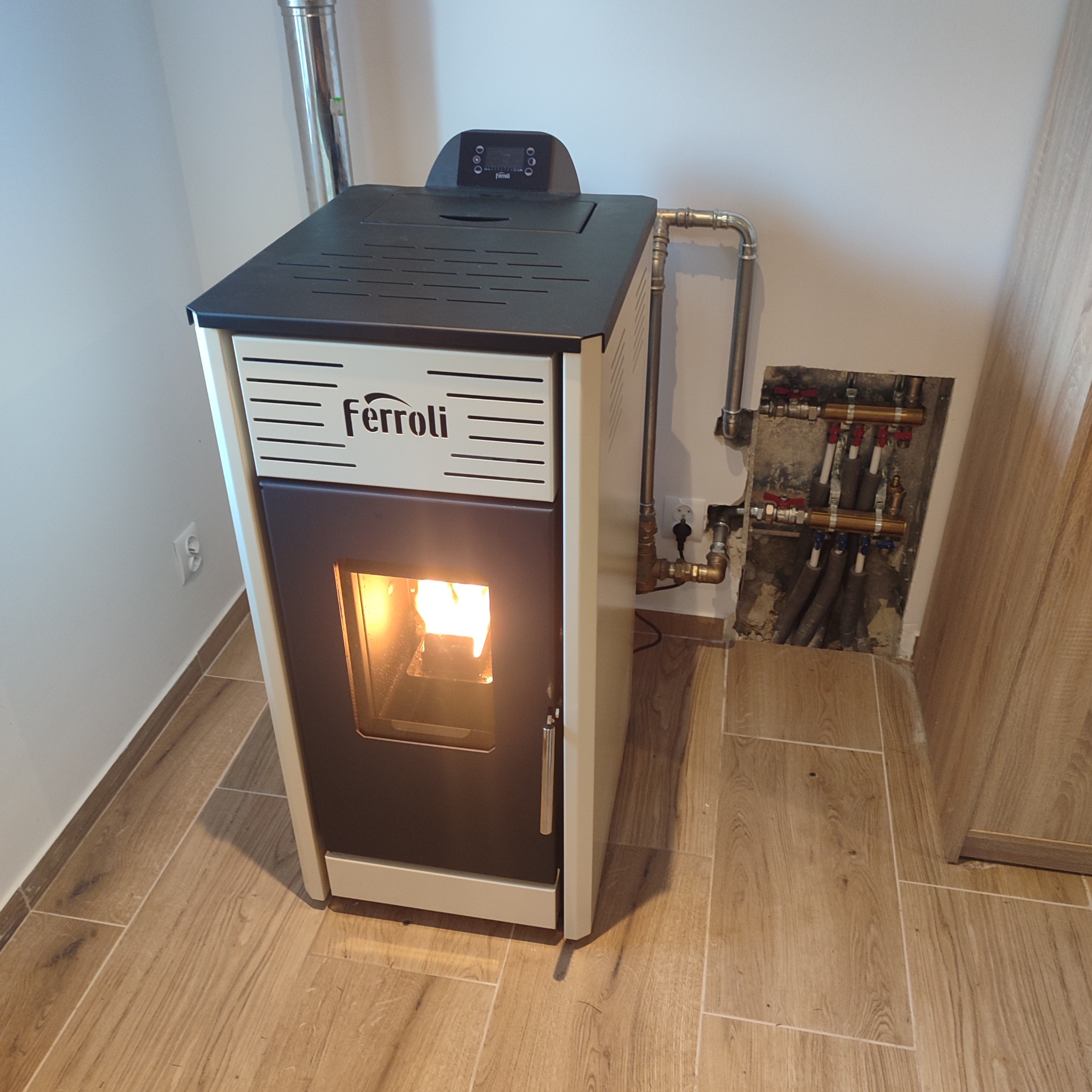 Palący się piec na pellet Ferroli z podłączeniem do instalacji grzewczej, widoczne rury i zawory w ścianie, drewnopodobne płytki na podłodze.
