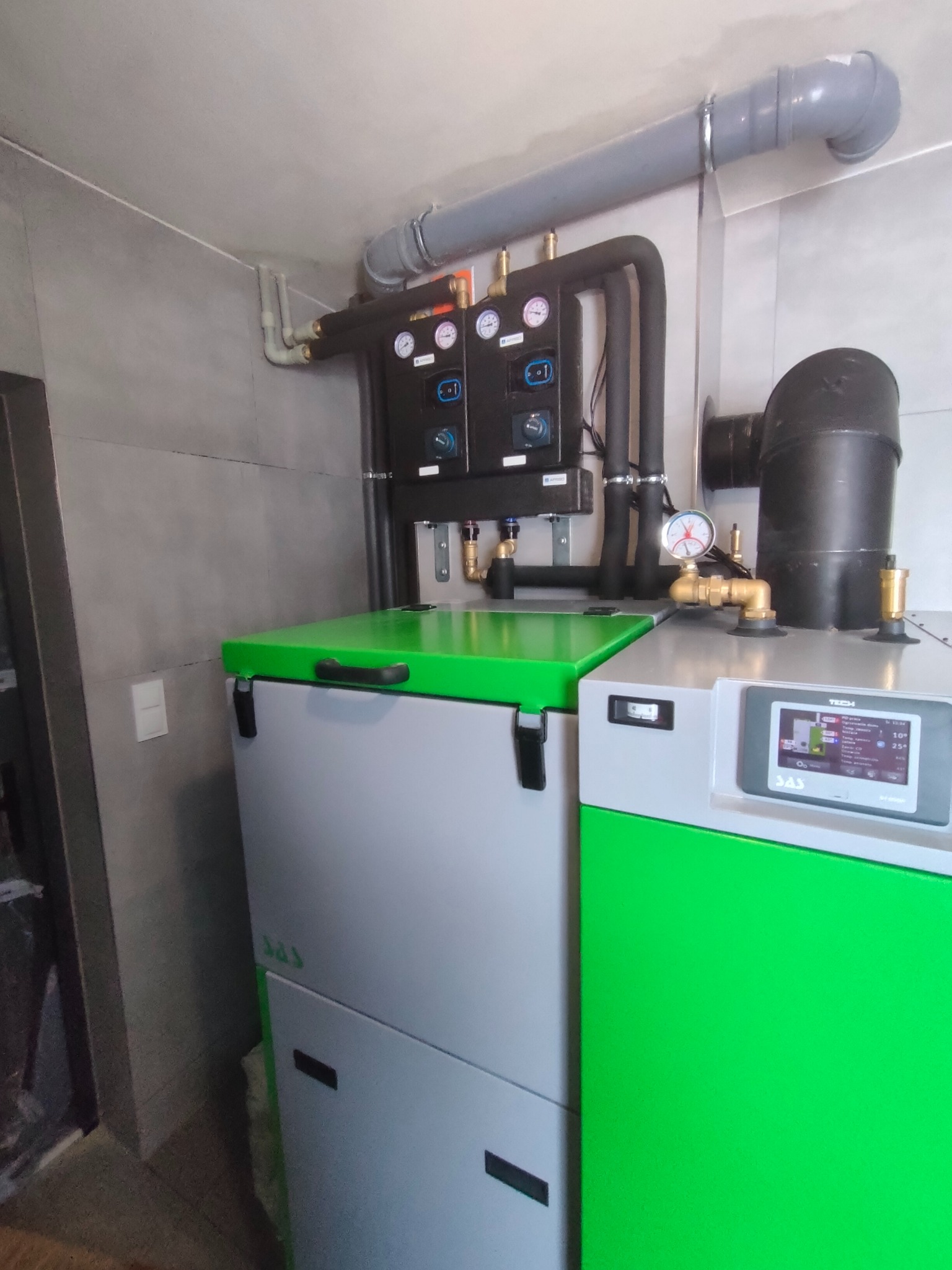 Nowoczesny piec CO z zielonym akcentem, zainstalowany w pomieszczeniu z szarą glazurą. Widoczne rury i czujniki, całość tworzy spójny system grzewczy.