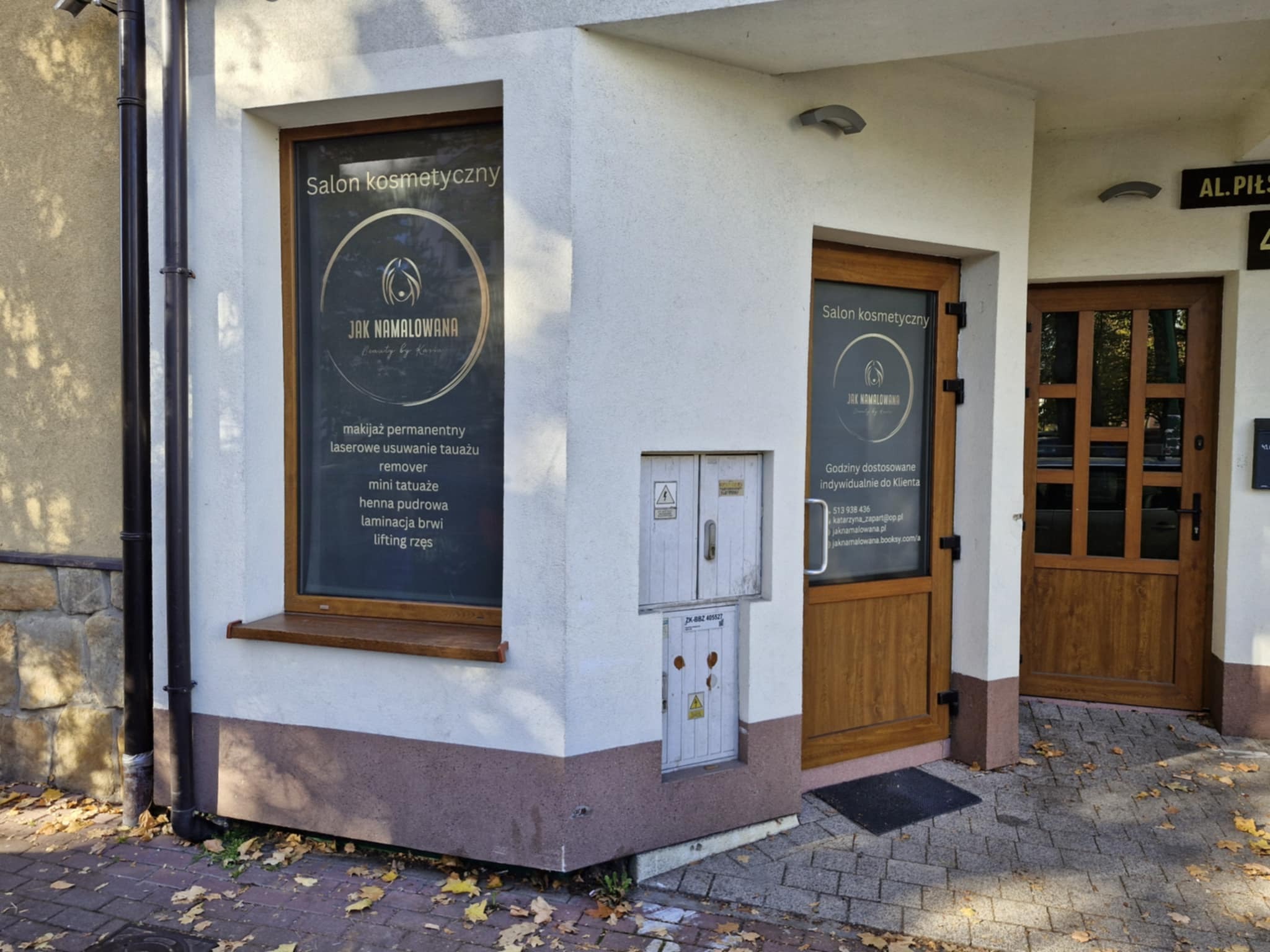 Wejście do salonu kosmetycznego z widocznymi na szybie informacjami o ofercie:  trwały, laserowe usuwanie tatuażu, mini tatuaże, henna pudrowa, laminacja brwi, lifting rzęs. Na drzwiach i oknie...