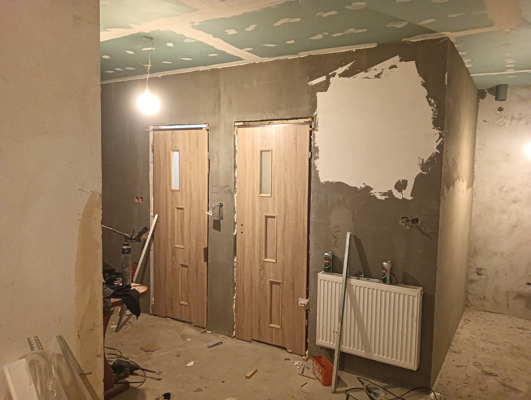 Wnętrze w trakcie remontu: dwie drewnopodobne drzwi z szybkami, surowa ściana z fragmentami gładzi, widoczne narzędzia i materiały budowlane, sufit w trakcie szpachlowania, grzejnik.