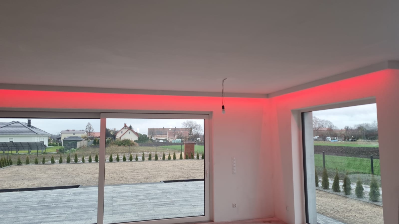 Wnętrze pokoju z oknem i przesuwnymi drzwiami balkonowymi, oświetlenie LED w kolorze czerwonym zamontowane wzdłuż sufitu, surowy stan wykończenia, widoczna żarówka na kablu.
