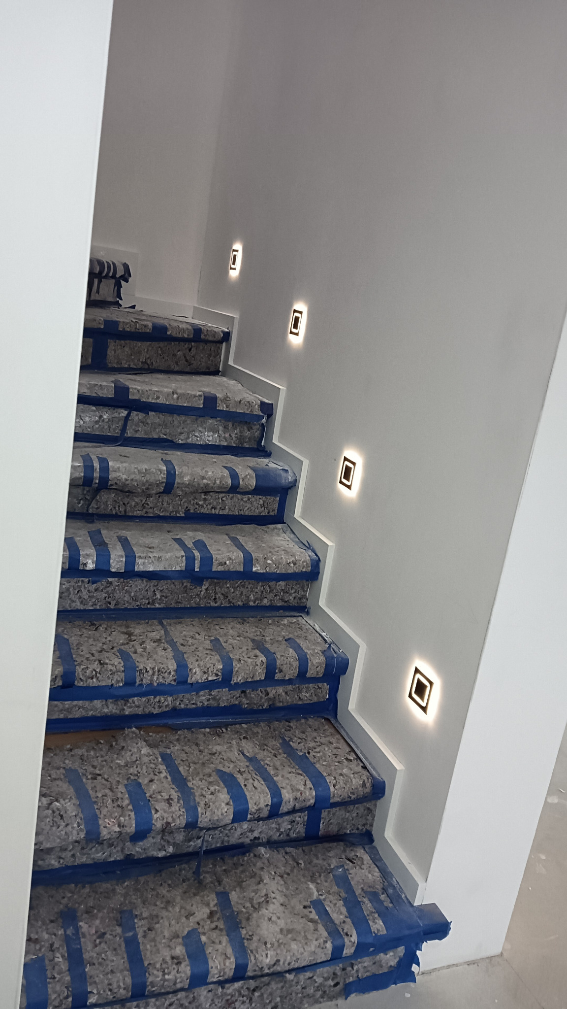 Schody zabezpieczone niebieską taśmą malarską przed uszkodzeniem, z zamontowanymi kwadratowymi lampami LED w ścianie.