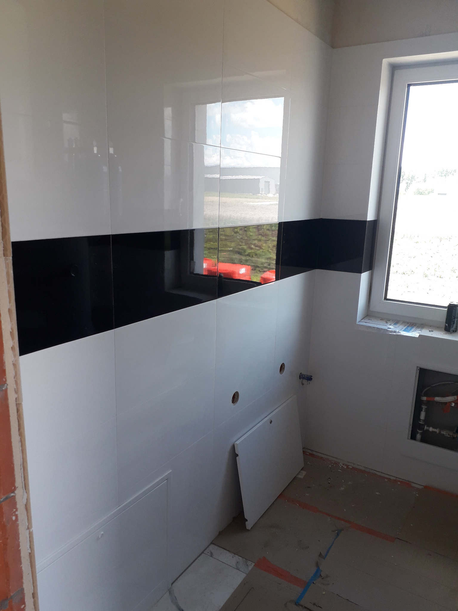 Wnętrze łazienki w trakcie prac wykończeniowych: białe płytki z poziomym pasem czarnych płytek, okno z widokiem na zewnątrz, widoczne instalacje wodne, fragment podłogi z resztkami kleju i taśmy...