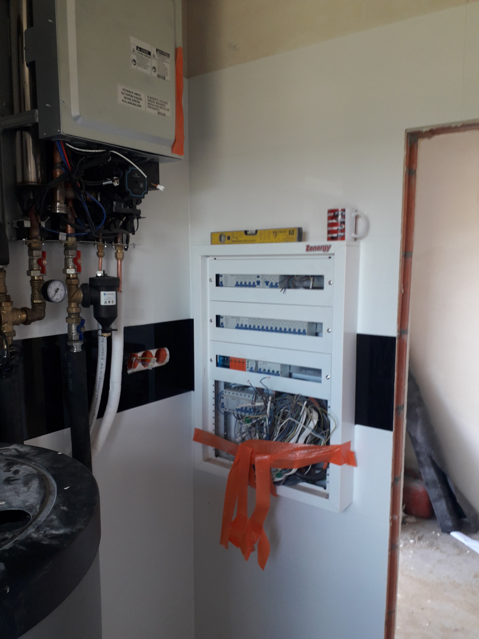 Wnętrze w trakcie prac: widoczny bojler, instalacja grzewcza z rurami miedzianymi i manometrem oraz otwarta skrzynka elektryczna z przewodami, poziomica na szafce, w tle fragment ściany z czarnymi...