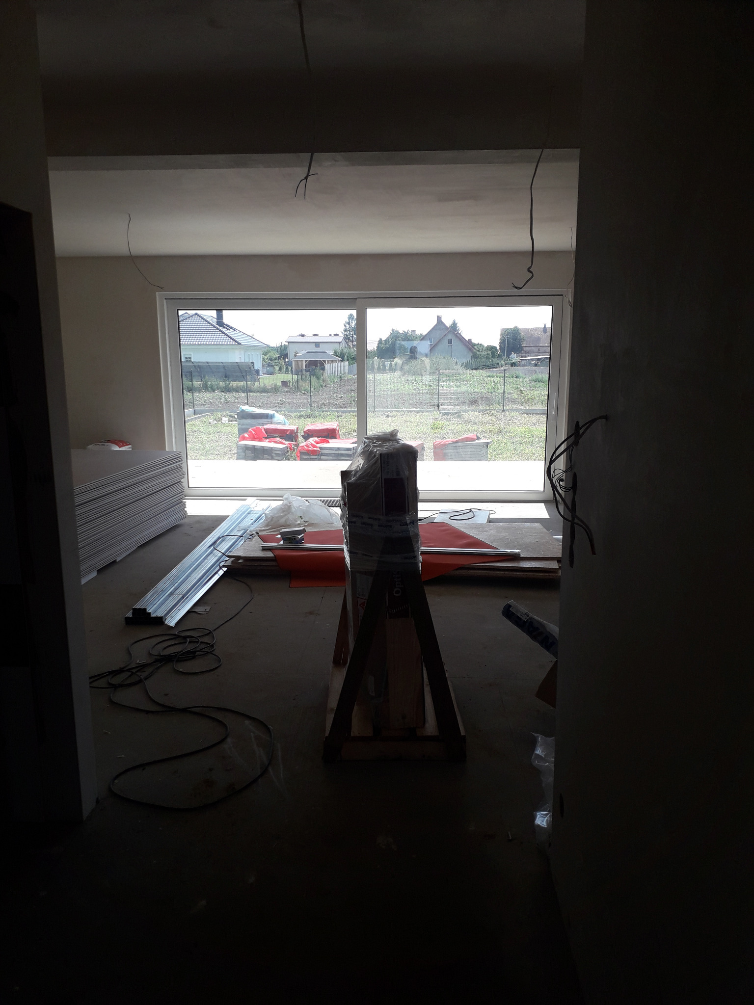 Wnętrze w trakcie prac wykończeniowych: widoczne płyty gipsowe, profile metalowe, narzędzia i materiały budowlane na surowej posadzce, z oknem na zewnątrz.