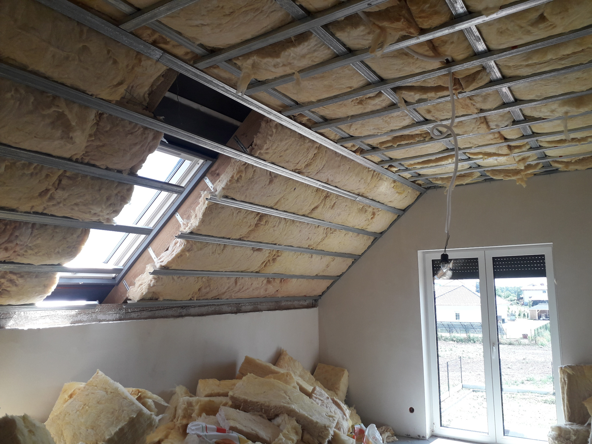 Montaż izolacji z wełny mineralnej w poddaszu, widoczna konstrukcja metalowa sufitu, okno dachowe i sterta materiału izolacyjnego na podłodze.