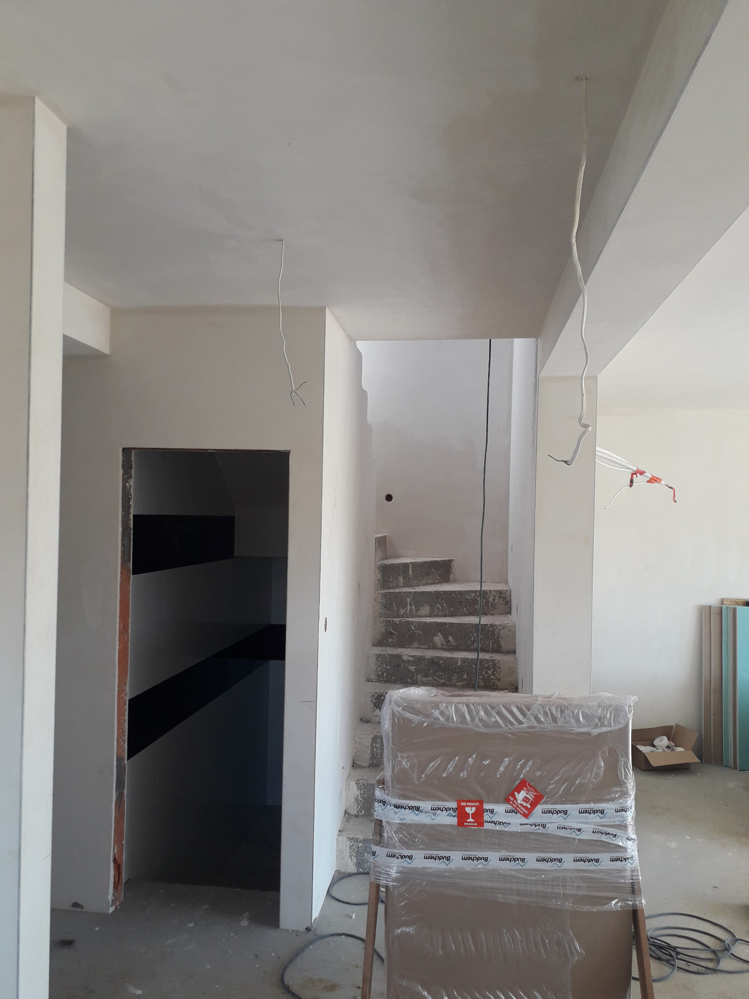 Surowe schody betonowe podczas prac budowlanych, widoczne przewody elektryczne zwisające z sufitu, ściany po wstępnym szpachlowaniu, zapakowane płyty gipsowe w folii z etykietą ostrzegawczą 'Fragile'.