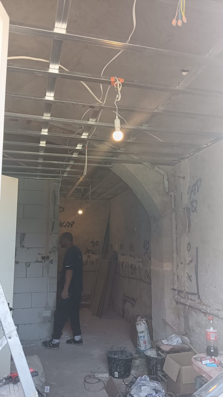 Wnętrze w trakcie remontu: widoczna konstrukcja sufitu podwieszanego z profili metalowych, instalacja elektryczna, ściany w stanie surowym z widocznymi napisami, pracownik w tle, bałagan...