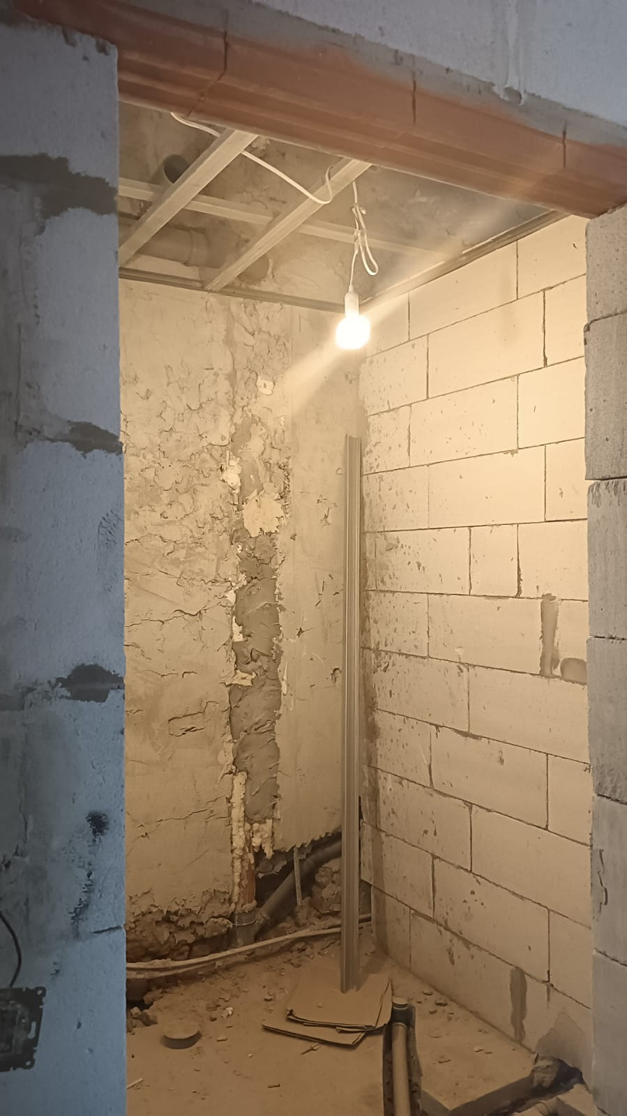 Wnętrze w trakcie prac przygotowawczych do wykończenia: ściana z bloków, fragment ściany ze starą farbą i resztkami zaprawy, wisząca żarówka, widoczne rury i kable, podłoga z resztkami gruzu.