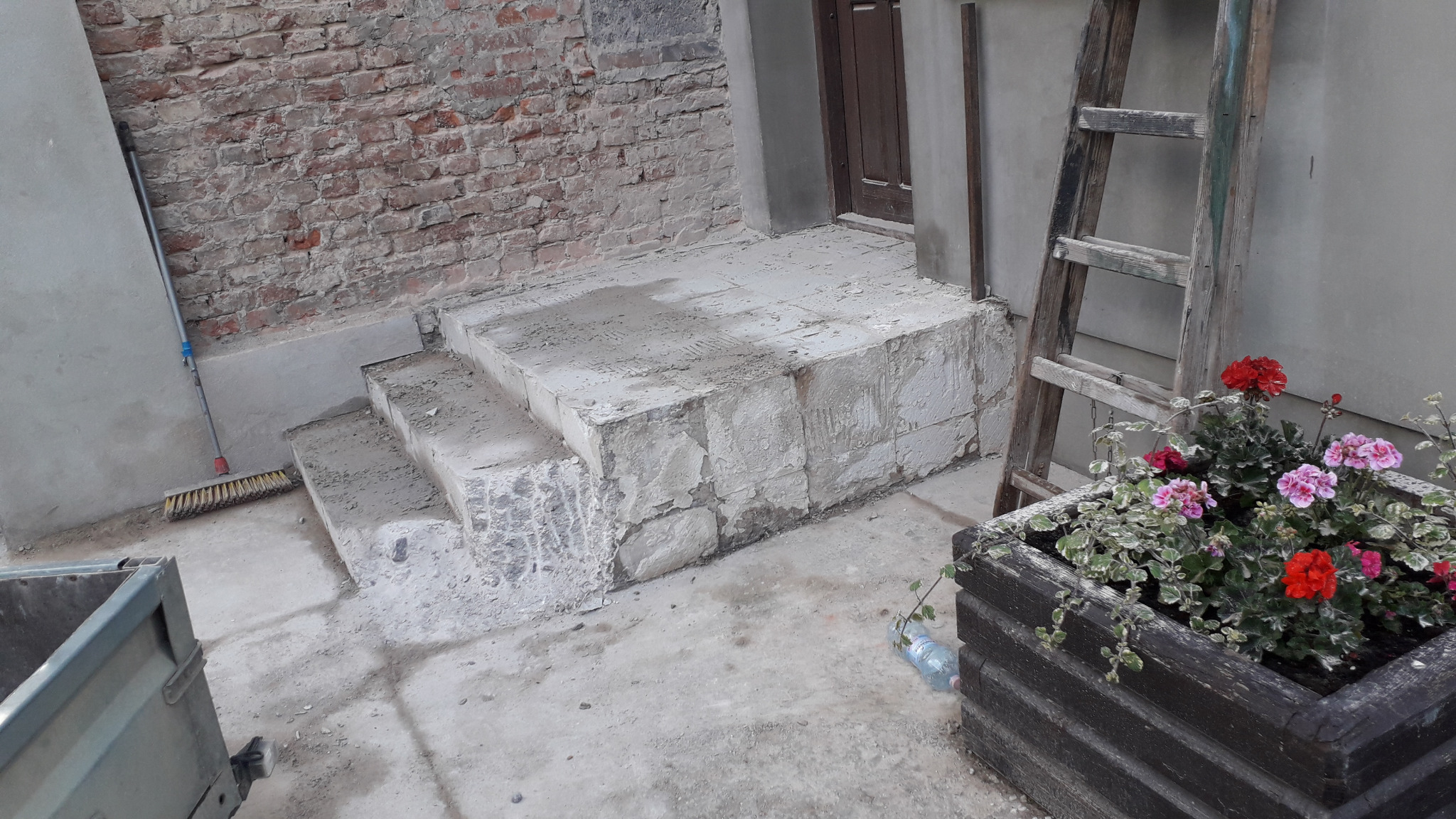 Betonowe schody wejściowe w trakcie budowy, z widocznym fragmentem ceglanej ściany, drabina drewniana oparta o ścianę i skrzynka z kwiatami.