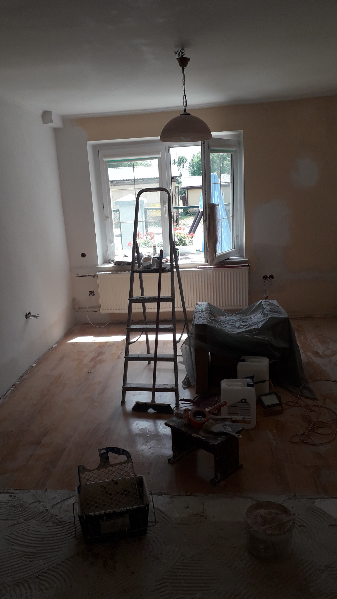 Pokój w trakcie prac wykończeniowych: drabina, narzędzia malarskie, częściowo pomalowane ściany, zabezpieczone meble i podłoga, widoczne puszki z farbą.