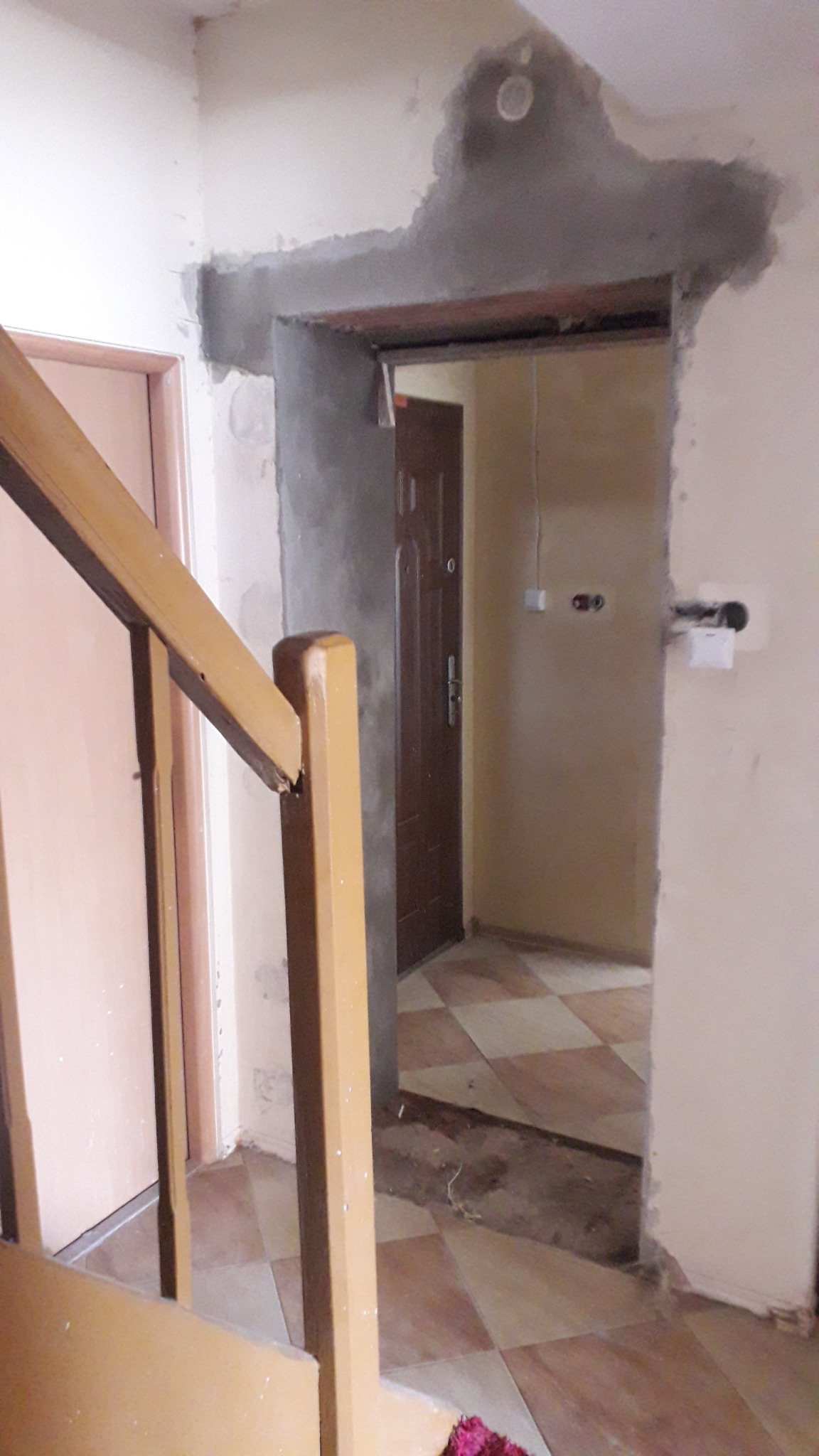 Niedokończone przejście drzwiowe z surowym betonem, widok zza drewnianej balustrady schodów, w tle brązowe drzwi wejściowe i podłoga wyłożona płytkami w romby.