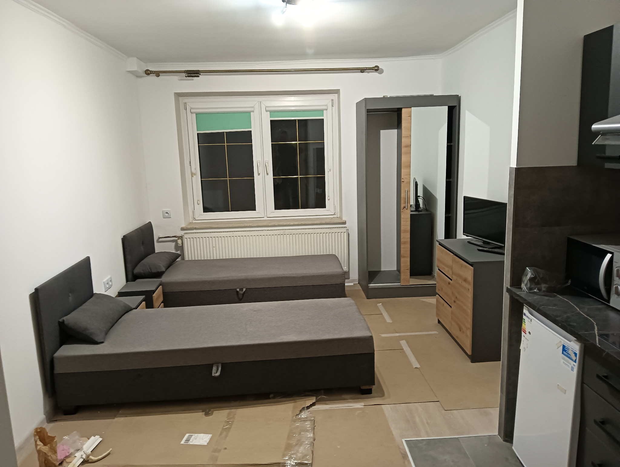 Pokój w trakcie wykończenia: dwa szare łóżka z zagłówkami, szafa z przesuwnymi drzwiami i telewizor na komodzie. Podłoga zabezpieczona kartonem, widoczny fragment aneksu kuchennego z lodówką...