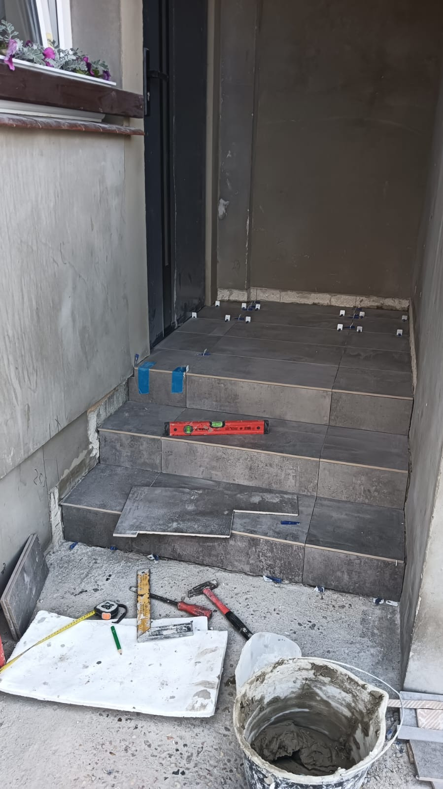 Kładzenie szarych płytek na schodach wejściowych, widoczne narzędzia: poziomica, młotek, miarka, kielnia, wiadro z zaprawą. Użyto krzyżyków dystansowych i taśmy malarskiej.
