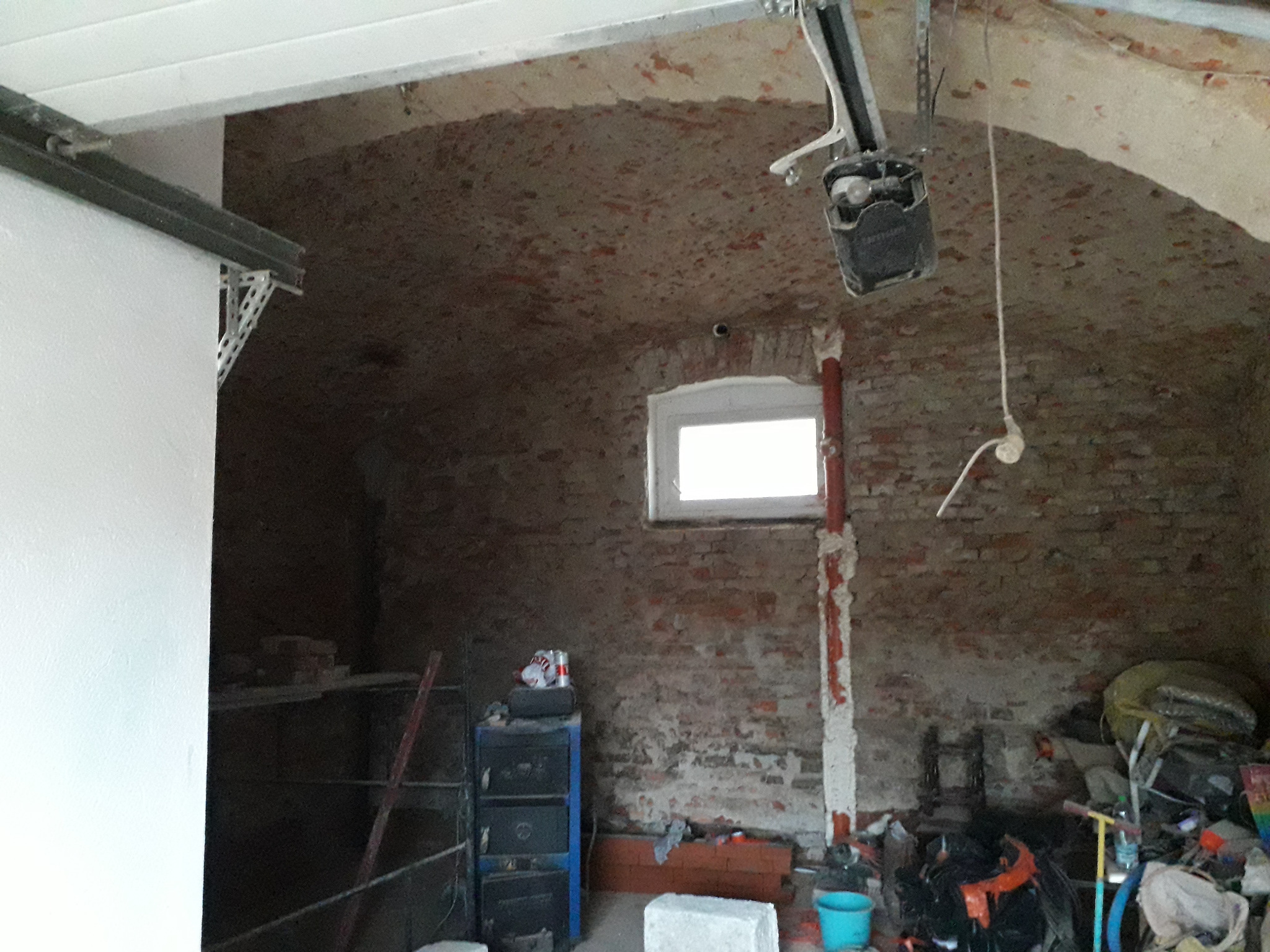 Wnętrze piwnicy ze sklepieniem łukowym z cegły, widoczne instalacje, okno i przedmioty składowane, ściana częściowo otynkowana i pomalowana na biało.