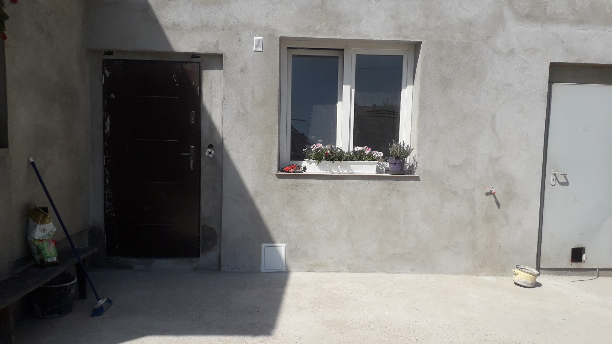 Surowa elewacja budynku z oknem, na parapecie doniczka z kwiatami, obok ciemne drzwi wejściowe i białe drzwi garażowe, widoczny cień, wiadro i miotła oparta o ławkę.