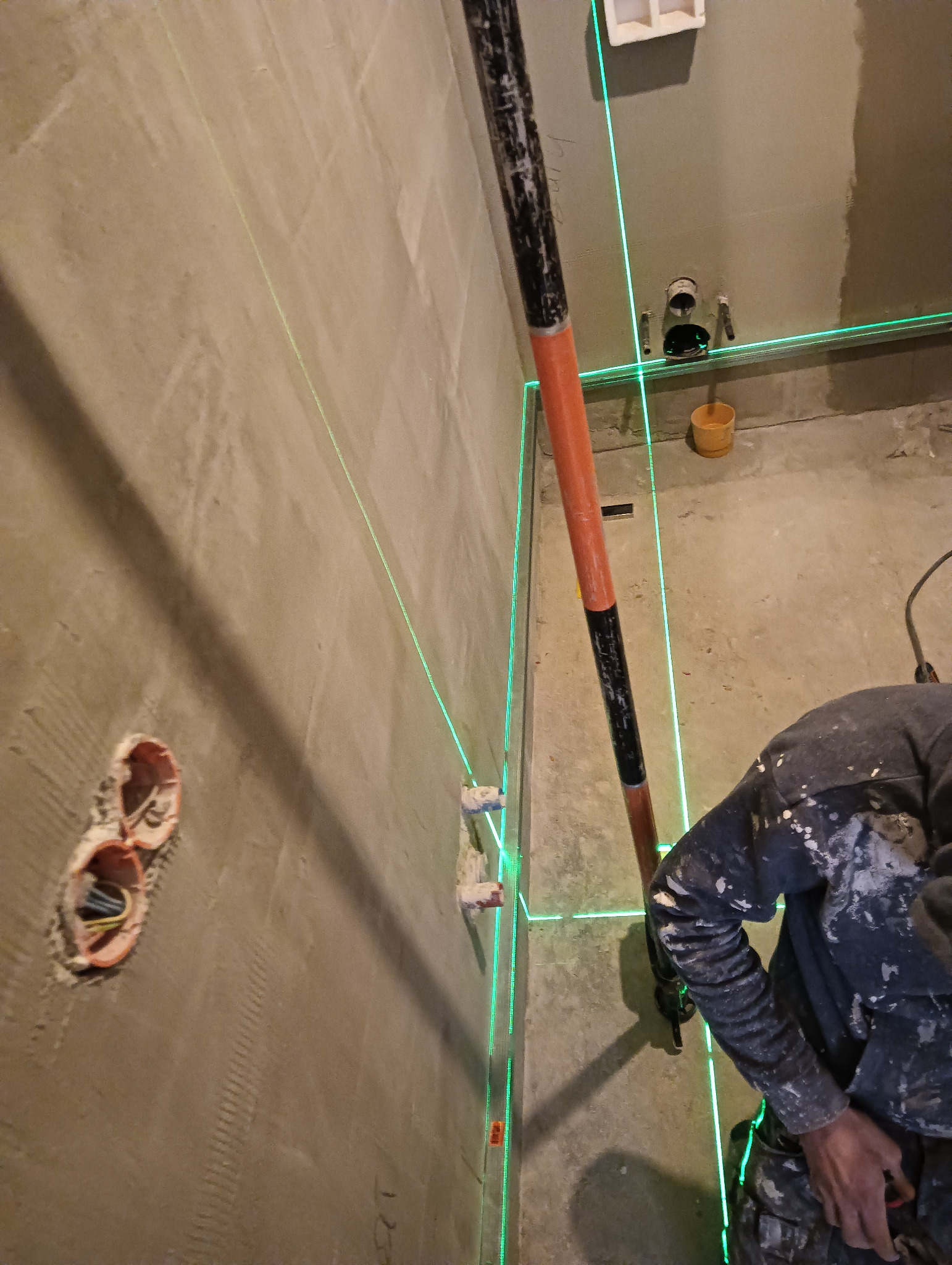 Ściana z nałożoną warstwą szpachli, widoczne puszki elektryczne, poziomica laserowa wyznacza linię, pracownik w ubraniu roboczym.